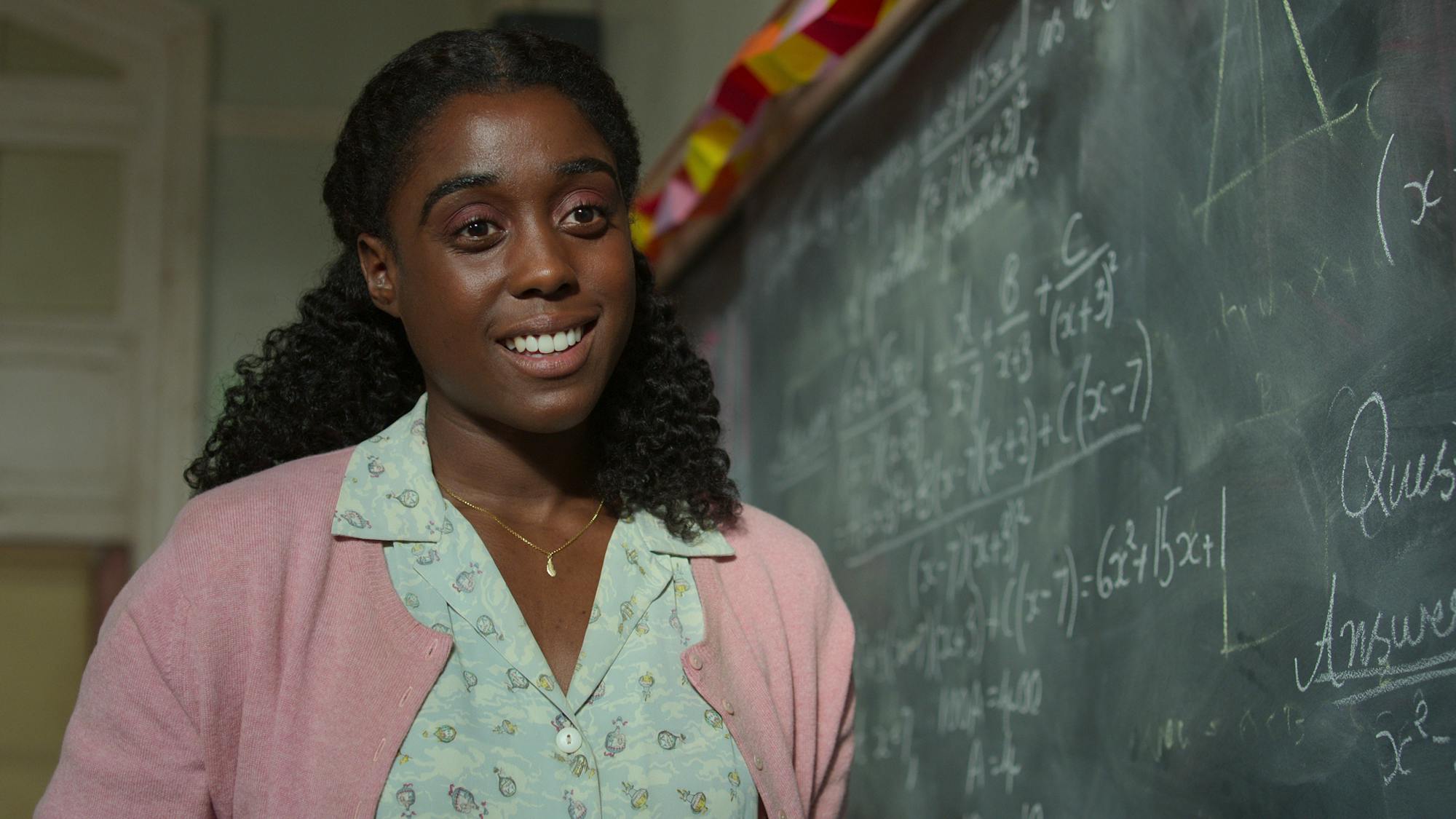 Miss Honey (Lashana Lynch) wears a green dress and a pink cardigan and stands next to a chalkboard.