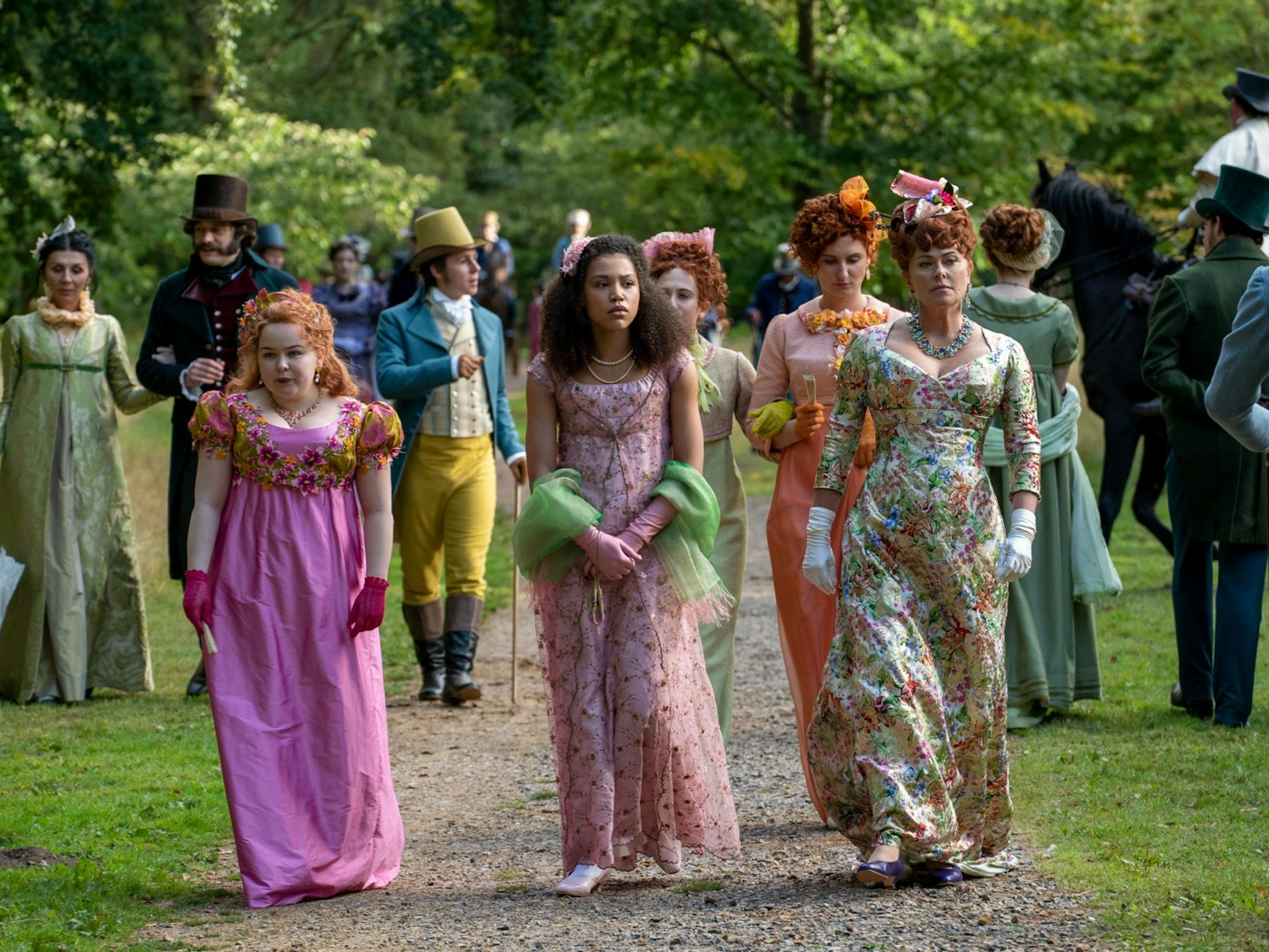 Penelope Featherington (Nicola Coughlan) and her family walk down a gravel path. They are surrounded by other people in equally as elaborate gowns, and men wearing suits with top hats. Between the prints and the greenery, the scene is bright and cheery despite Lady Featherington’s cold expression.