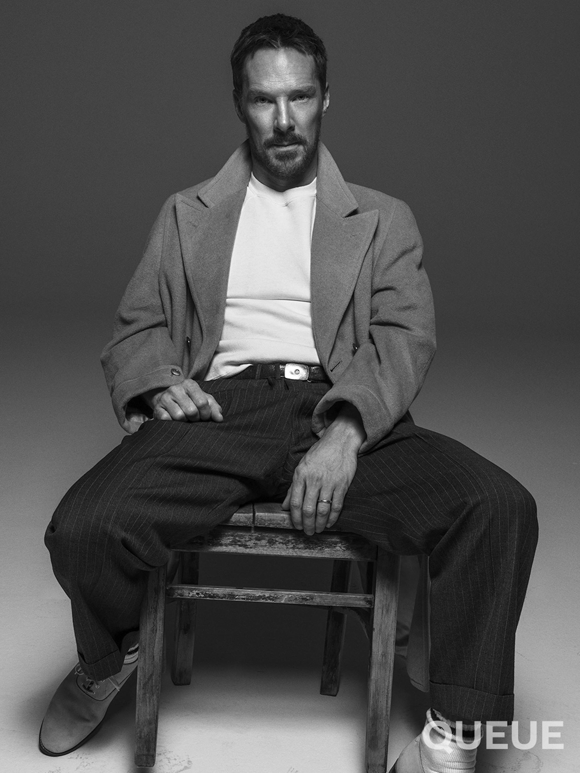 Benedict Cumberbatch wears dark pants, a white shirt, and a grey cardigan. He sits in a chair looking straight at the camera.
