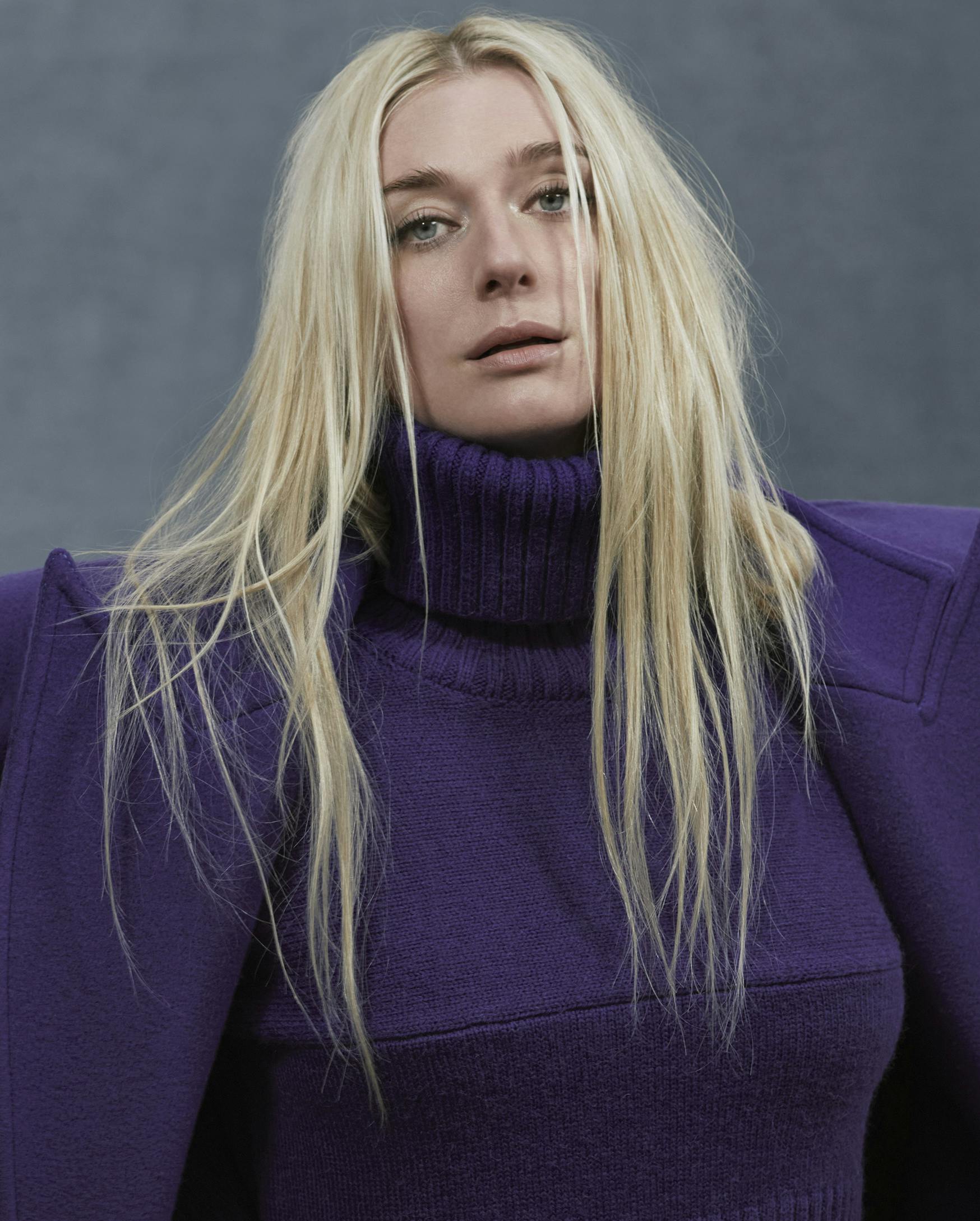 Elizabeth Debicki wears a purple sweater, coat, and pants and stands against a gray wall. 