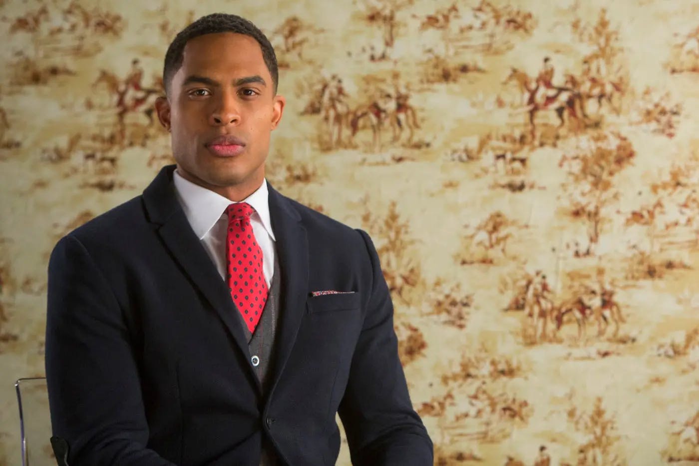 Troy Fairbanks (Brandon Bell) sits in front of a vintage patterned background of horses & their owners.  He looks confident and serious and is smartly dressed in a navy blue suit, grey waistcoat, a red tie with green dots, and a patterned pocket-handkerchief.
