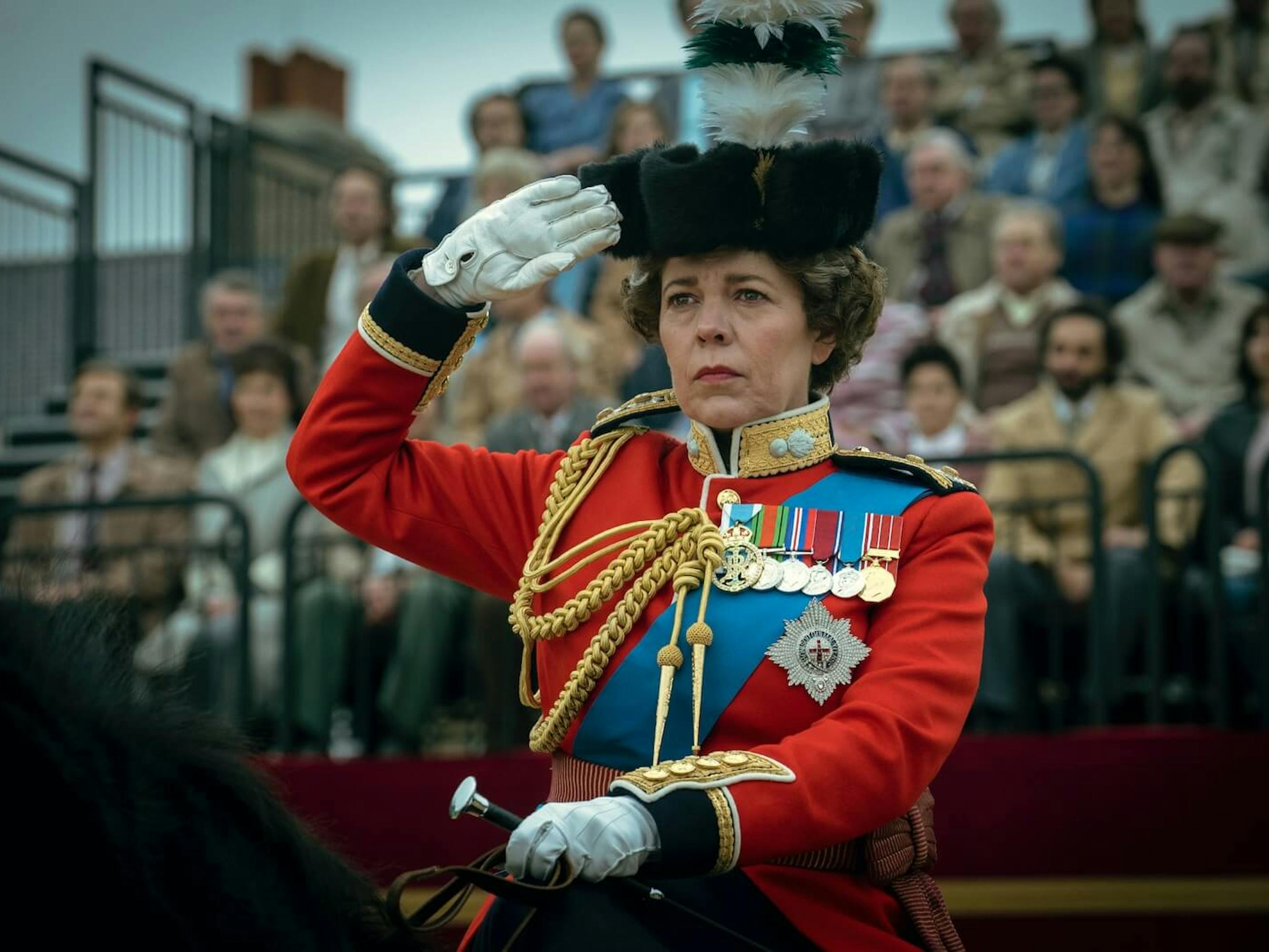 Dressed in adorned red military garb, Queen Elizabeth (Olivia Colman) salutes a crowd from atop a horse.