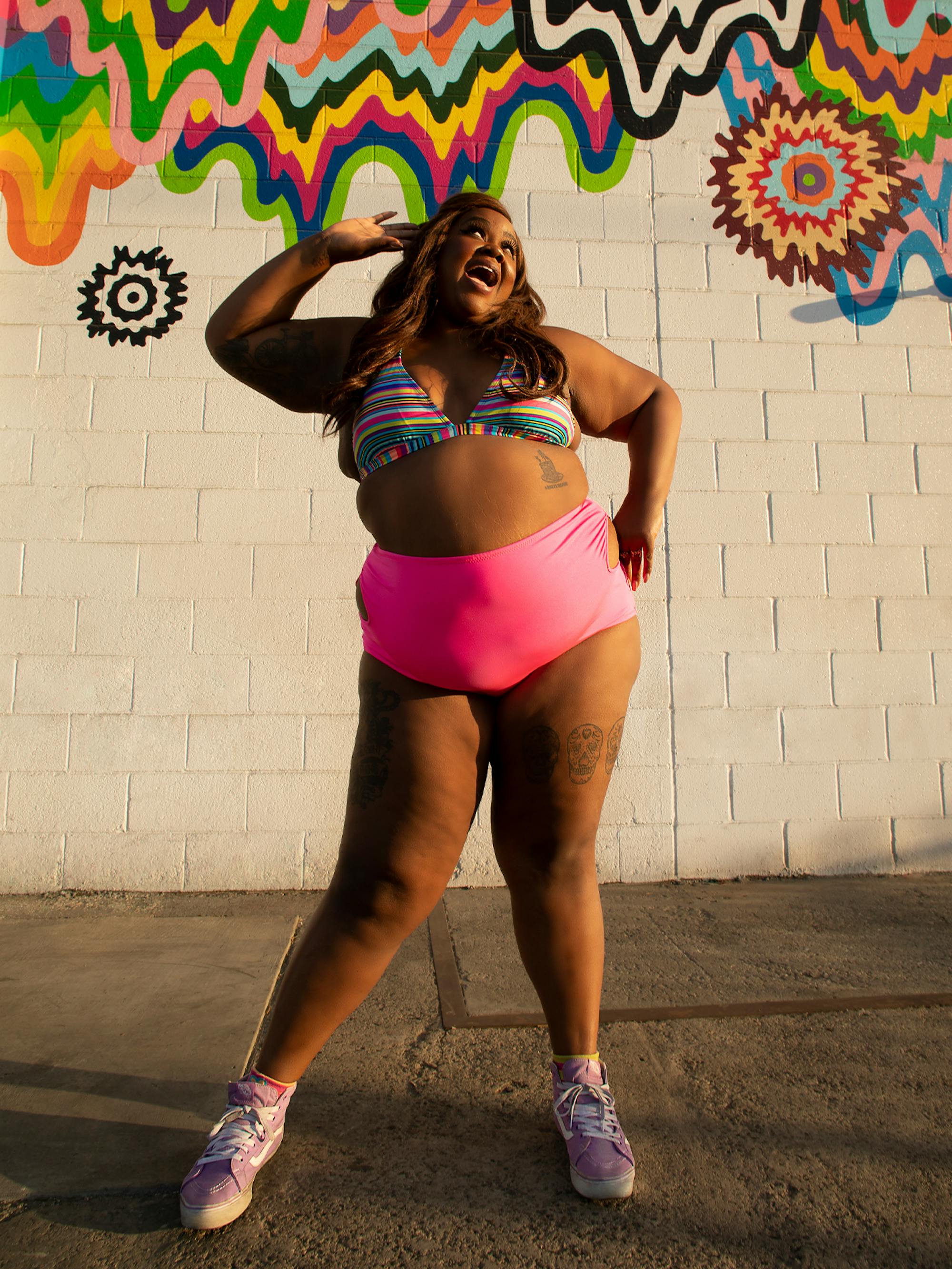 Nicole Byer poses in front of a sunlit graffiti wall. She wears a bikini with bright pink pants and trainers.