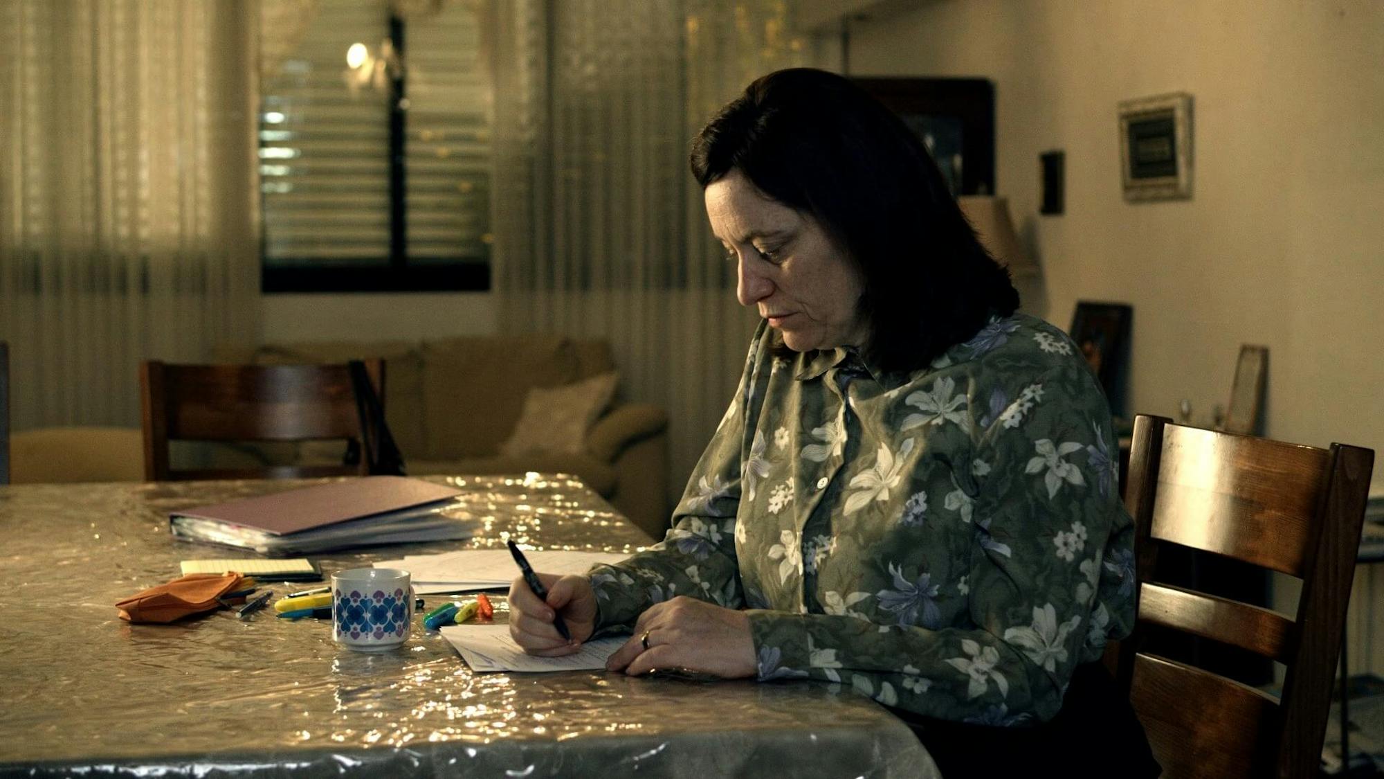 Tovi Shtisel (Eliana Shechter) sits at a plastic wrapped table writing something. She wears a green patterned top. The room is dark.