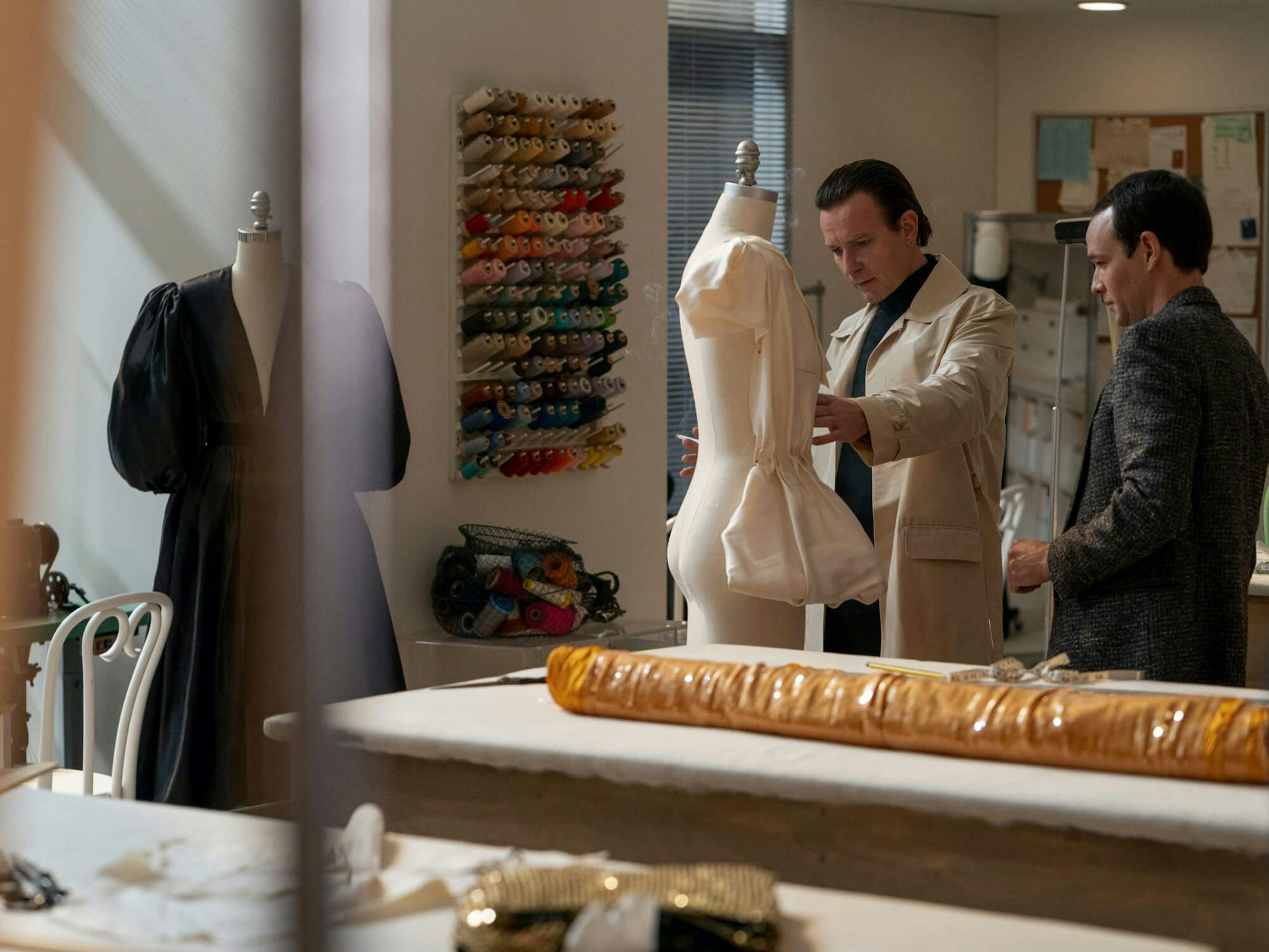 Halston examines a white dress on a mannequin in his studio, accompanied by a man in a dark blazer.
