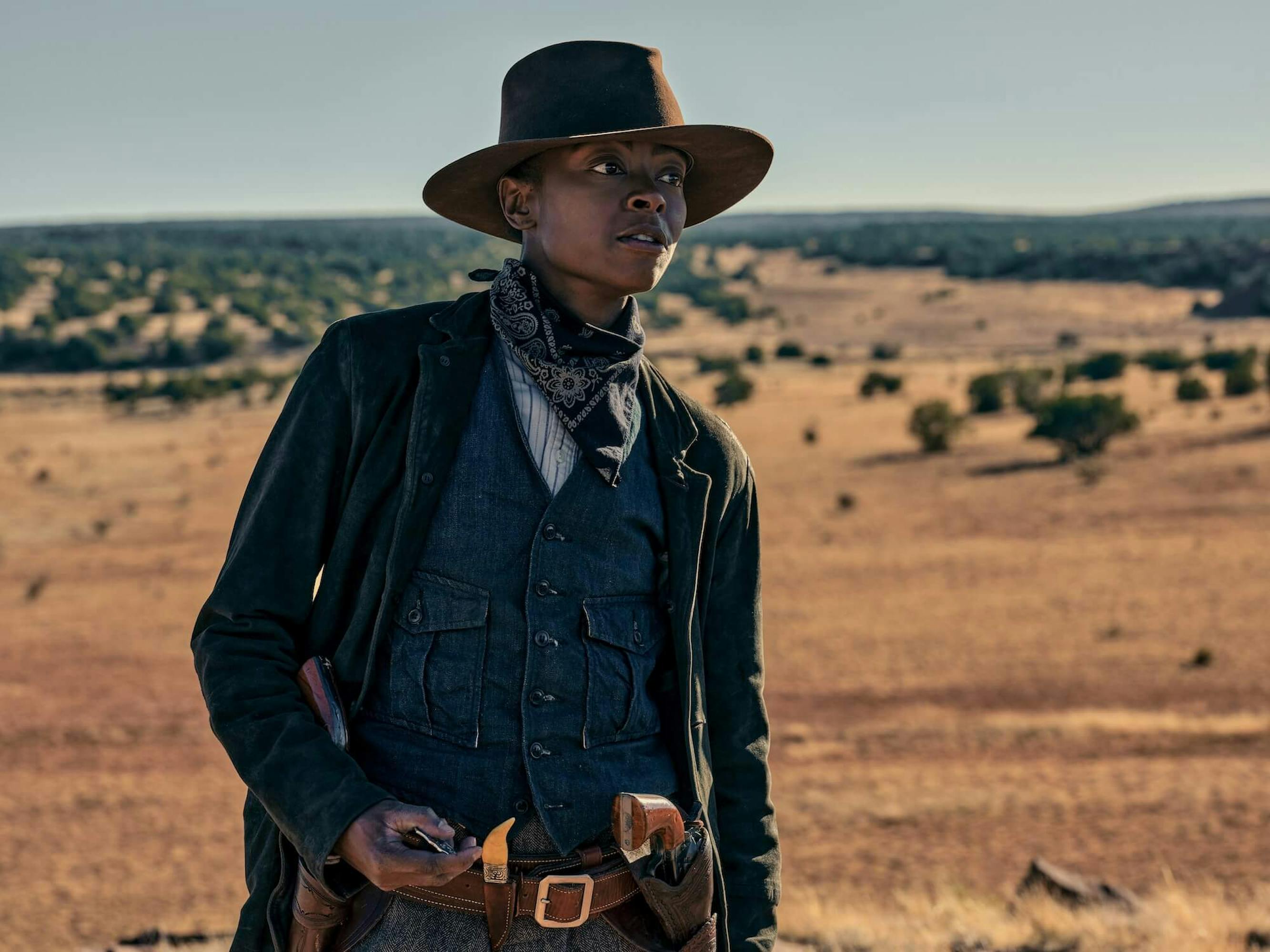 Cuffee (Danielle Deadwyler) wears a denim vest, green jacket, handkerchief, and a brown hat. Her belt is hooked with knives and guns.