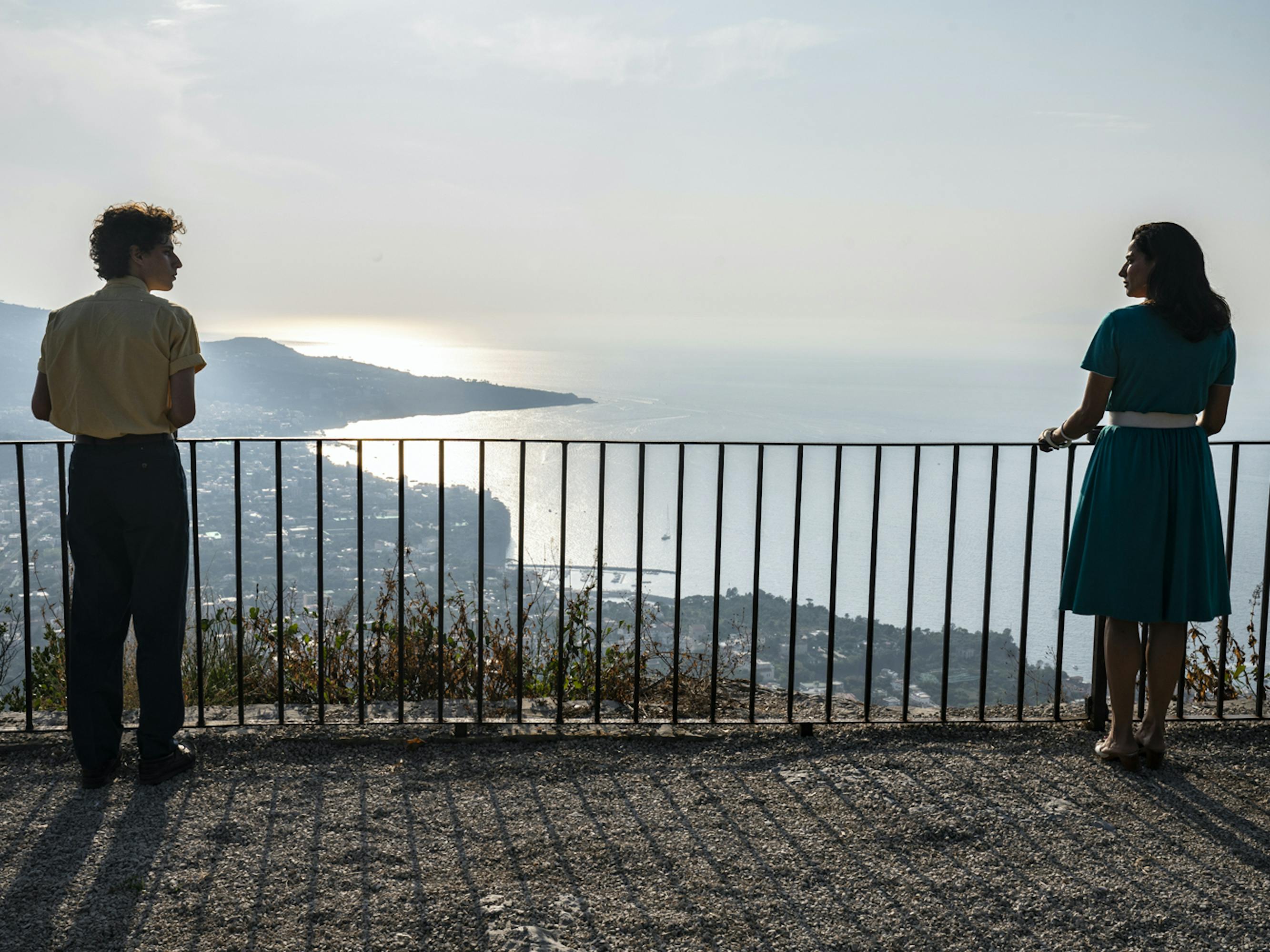 Fabietto Schisa (Filippo Scotti) and Patrizia (Luisa Ranieri) stand beside a railing looking at each other and at the sunlit water. 