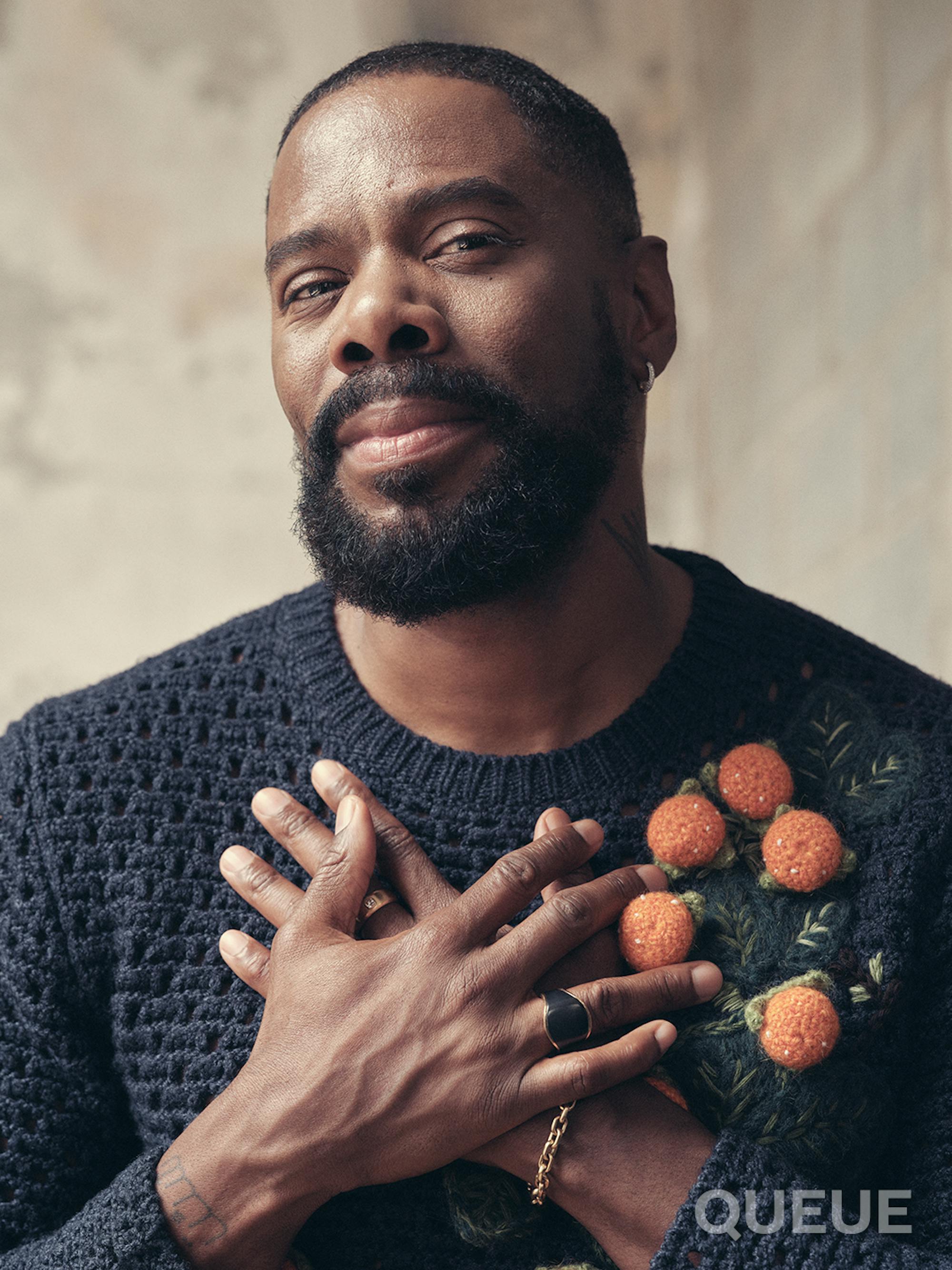 Colman Domingo holds his hands to his chest. He wears a navy blue sweater with 3D oranges on them. 