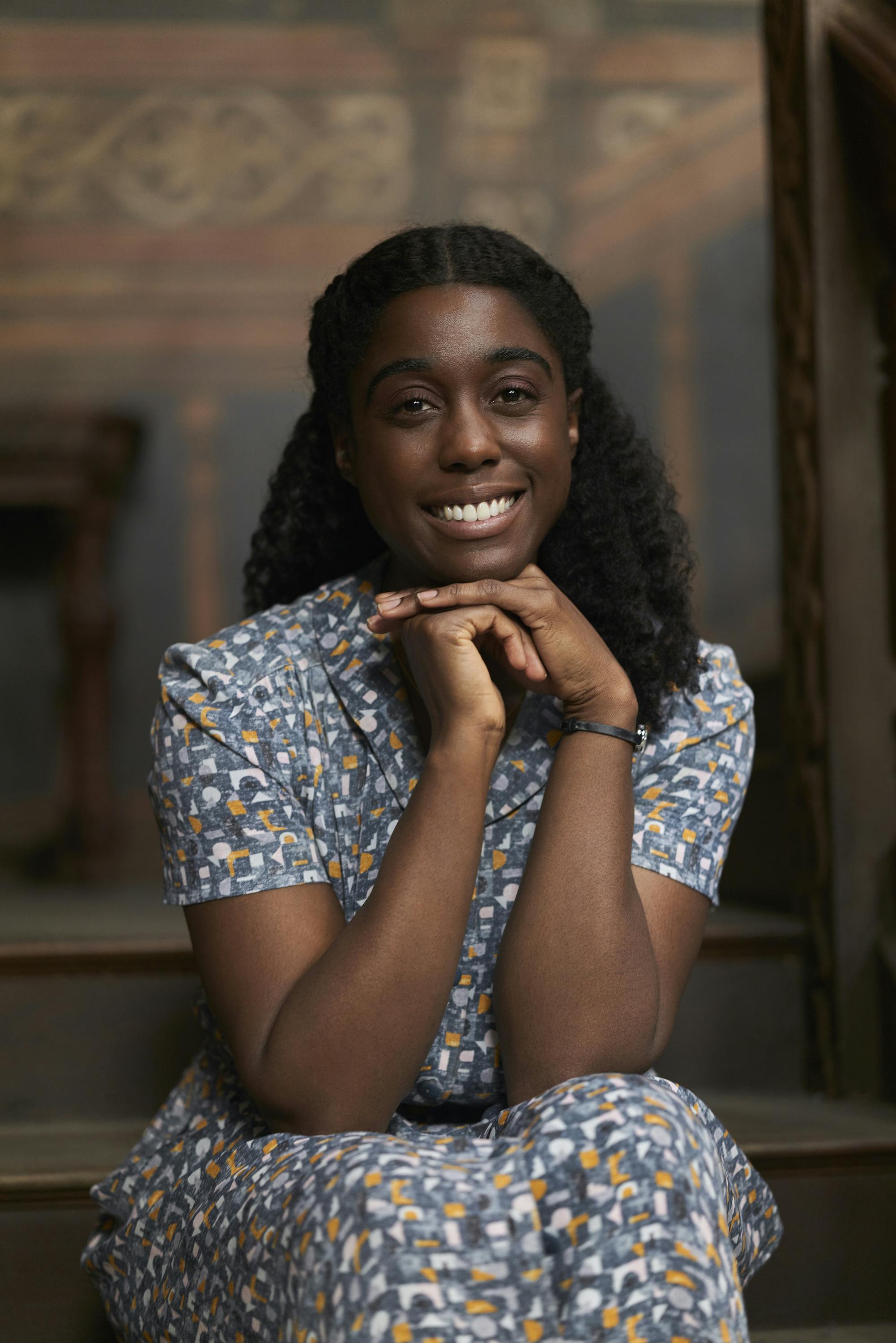 Lashana Lynch wears a patterned dress and rests her chin on her hands.