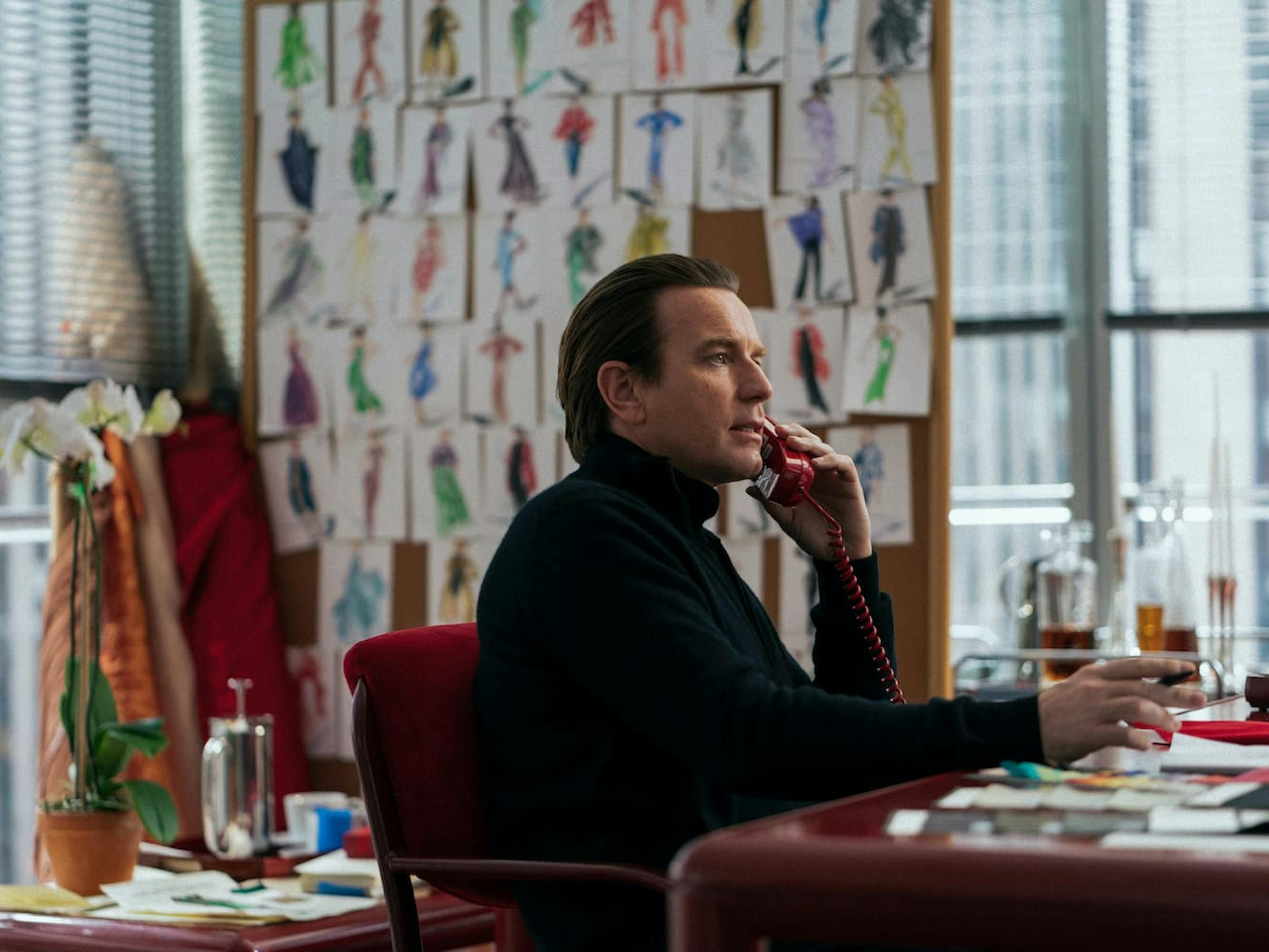 Halston (Ewan McGregor) wears a black turtleneck in an open office. In the background are sketches of dresses. He sits at a red desk, speaking into a red phone.