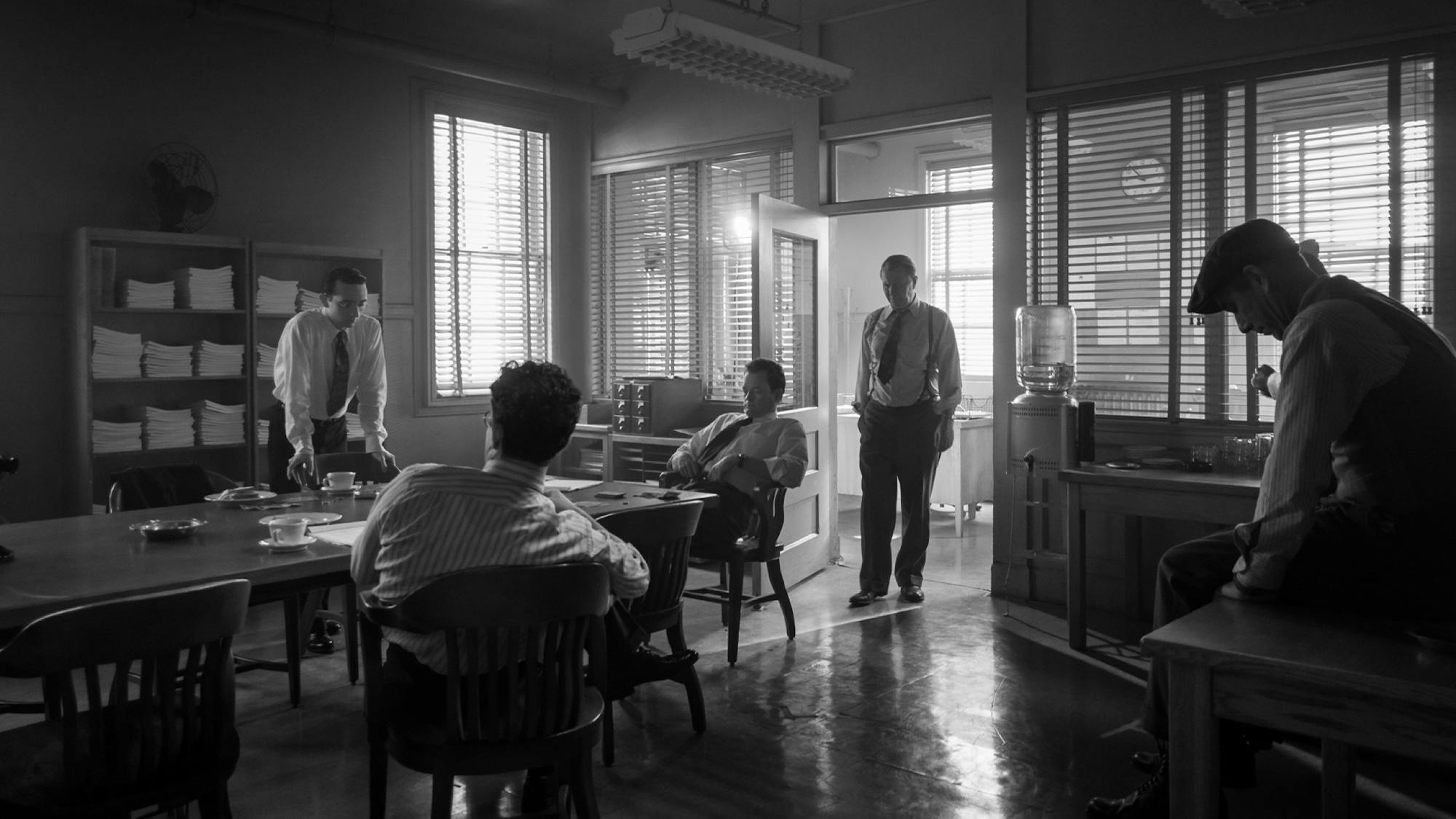  Irving Thalberg’s office in Mank. Venetian blinds filter the light into shafts. Men are perched throughout the office, some around a large conference table. Mank stands posed in a doorway. 