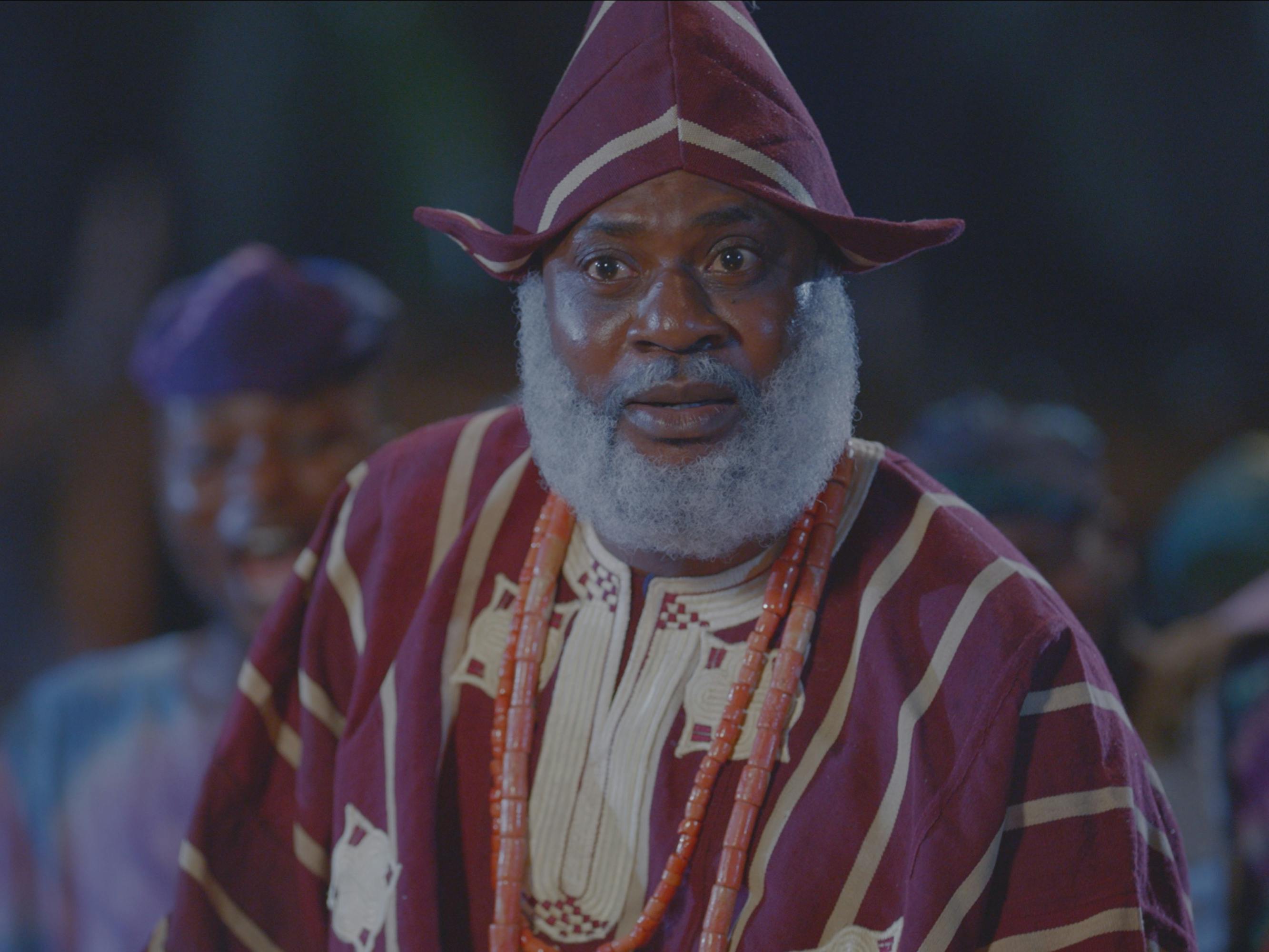 Elesin Oba (Odunlade Adekola) wears a red robe and hat and stands in dusky light.
