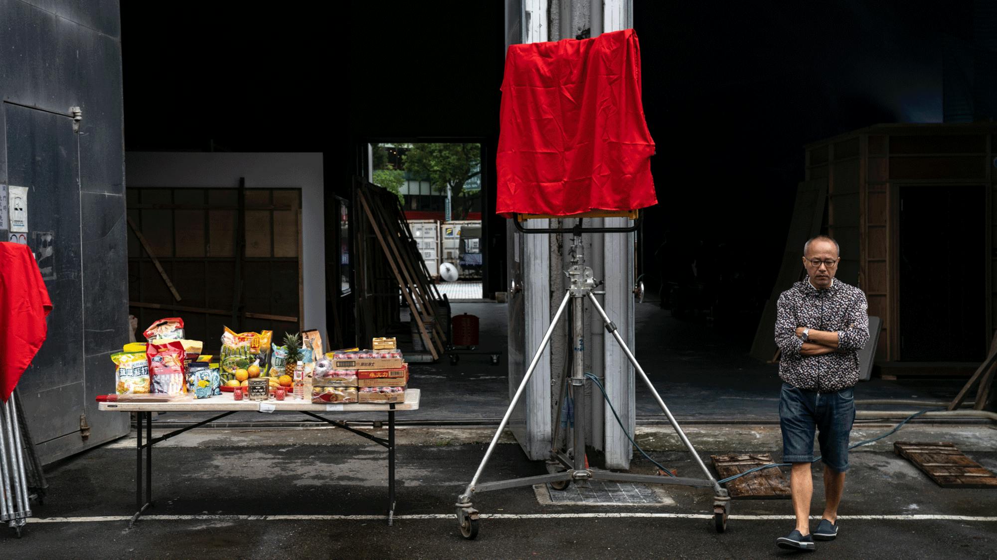 Director Chung Mong-hong is photographed behind the scenes on the set of A Sun. He stands next to a table of food in shorts and a sweater with his arms crossed over his chest.