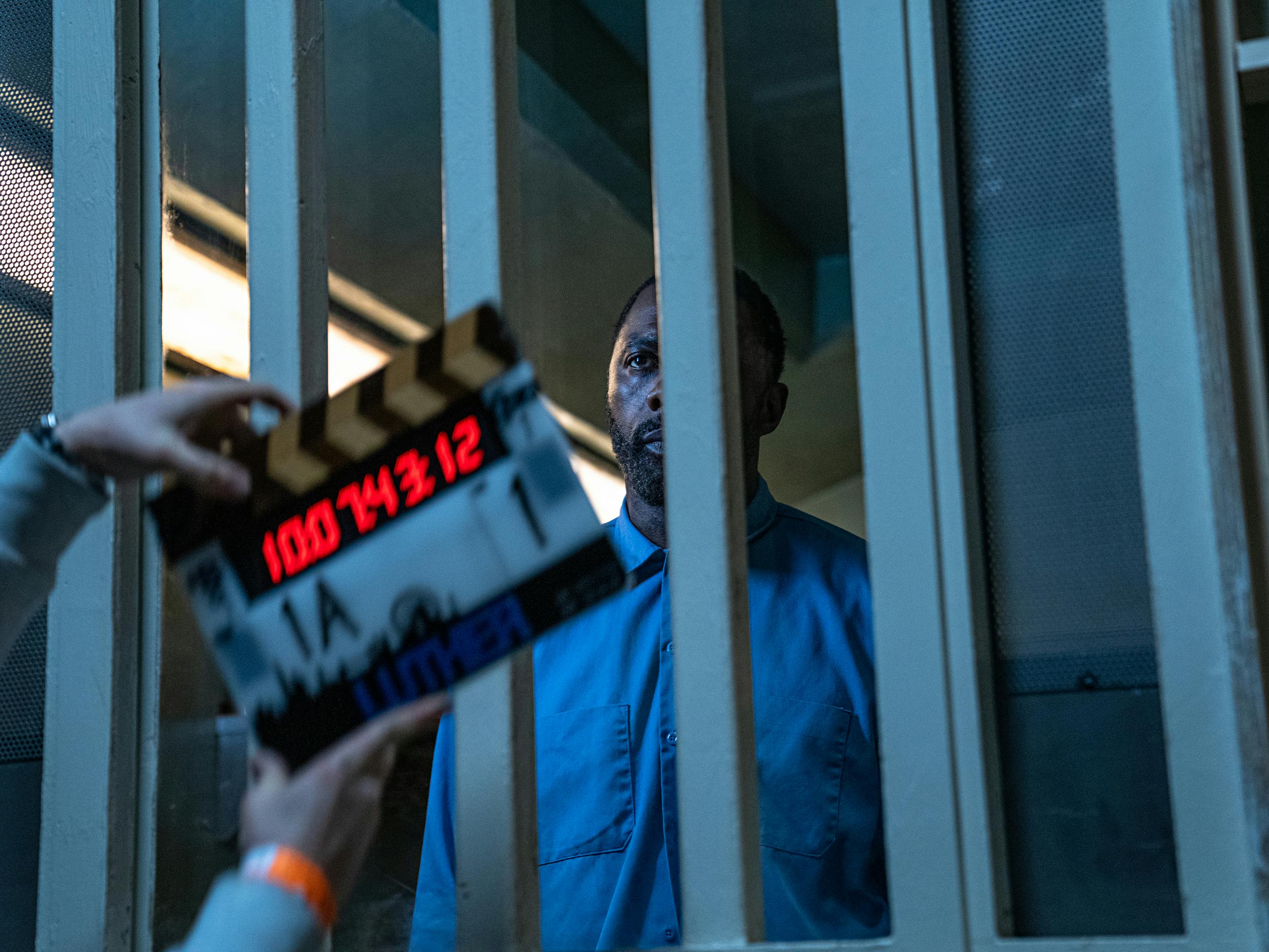 John Luther (Idris Elba) behind the scenes. He wears blue prison garb and looks intense. Partially covering his face is a clapperboard.
