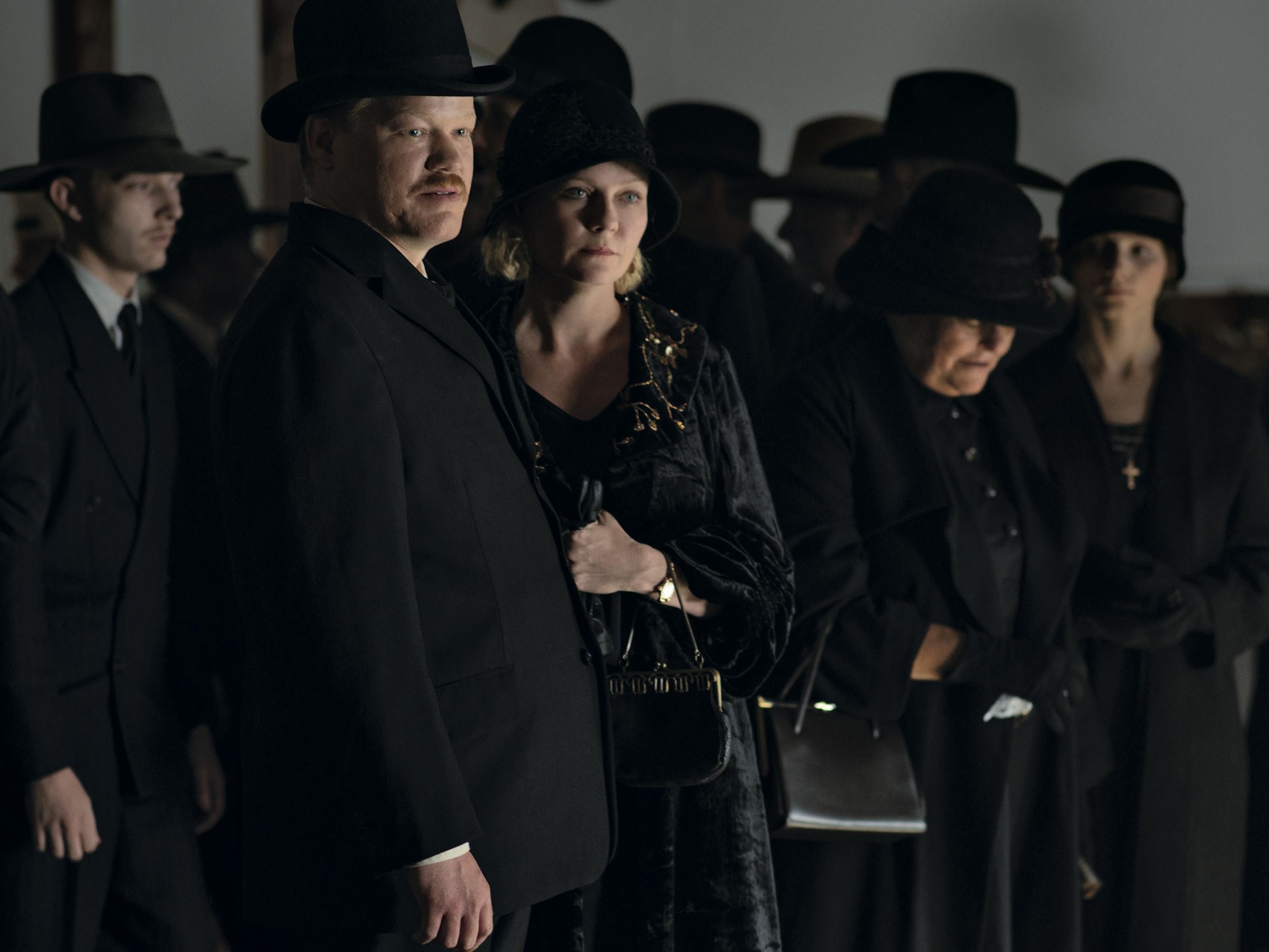 Jesse Plemons and Kirsten Dunst wear black and stand in a crowd of other people wearing black outfits and black hats.