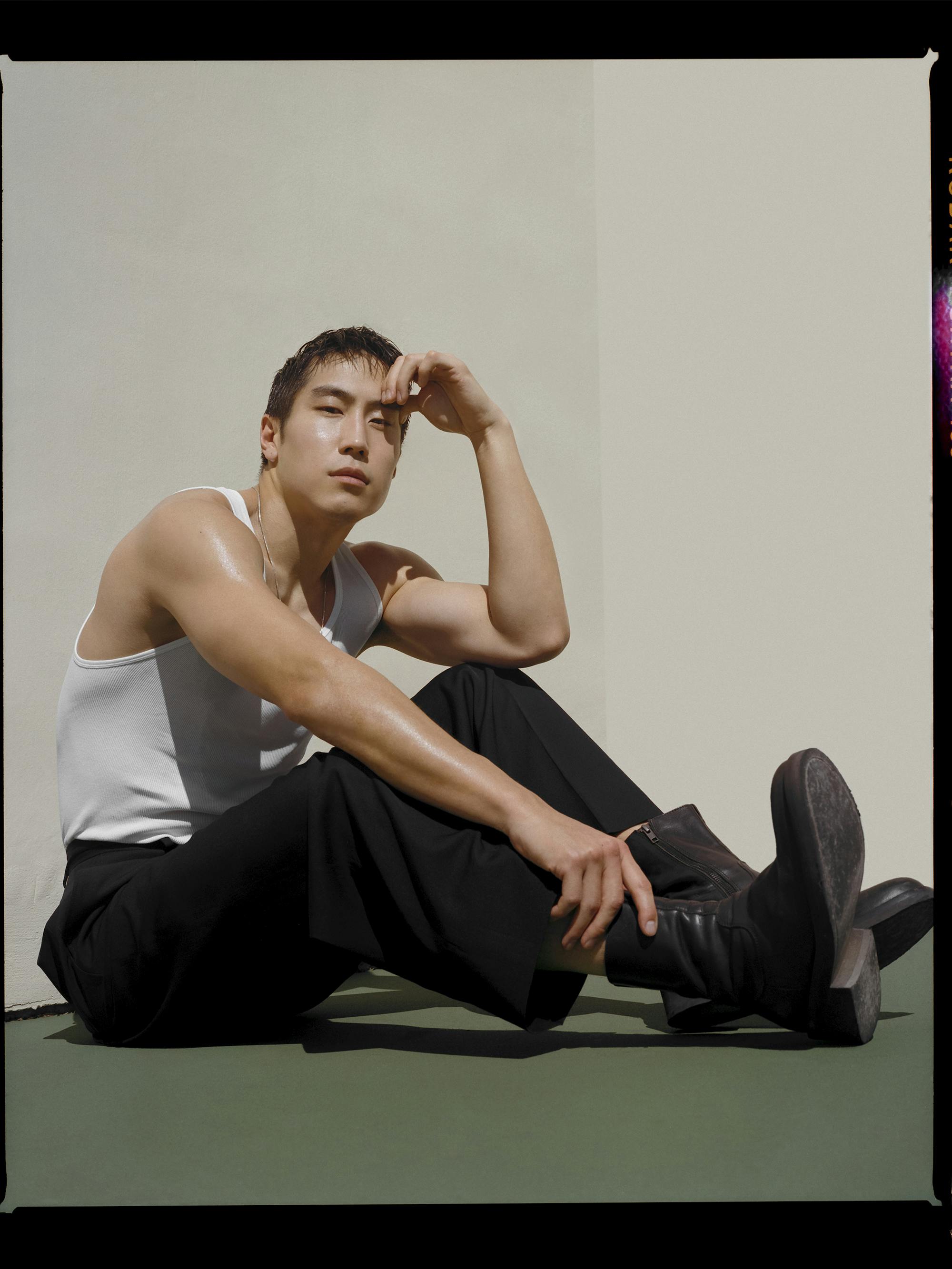 Young Mazino wears black pants and a white shirt and sits on a green floor.