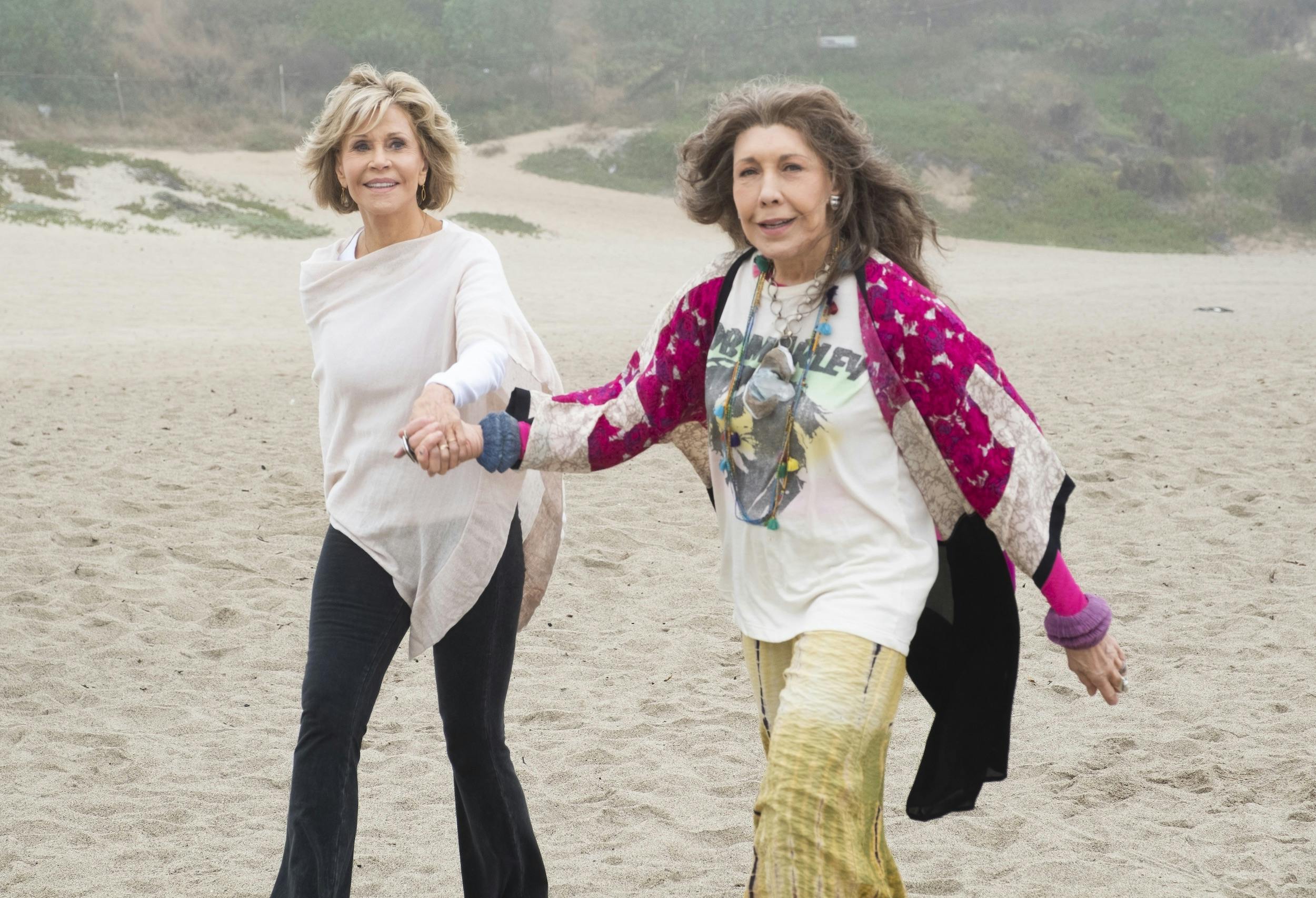 Grace and Frankie walk on a beach holding hands. Grace wears dark pants and a tan poncho. Frankie wears yellow pants, a white graphic tee, necklaces, and a pink wrap. They look happy in this foggy scene.