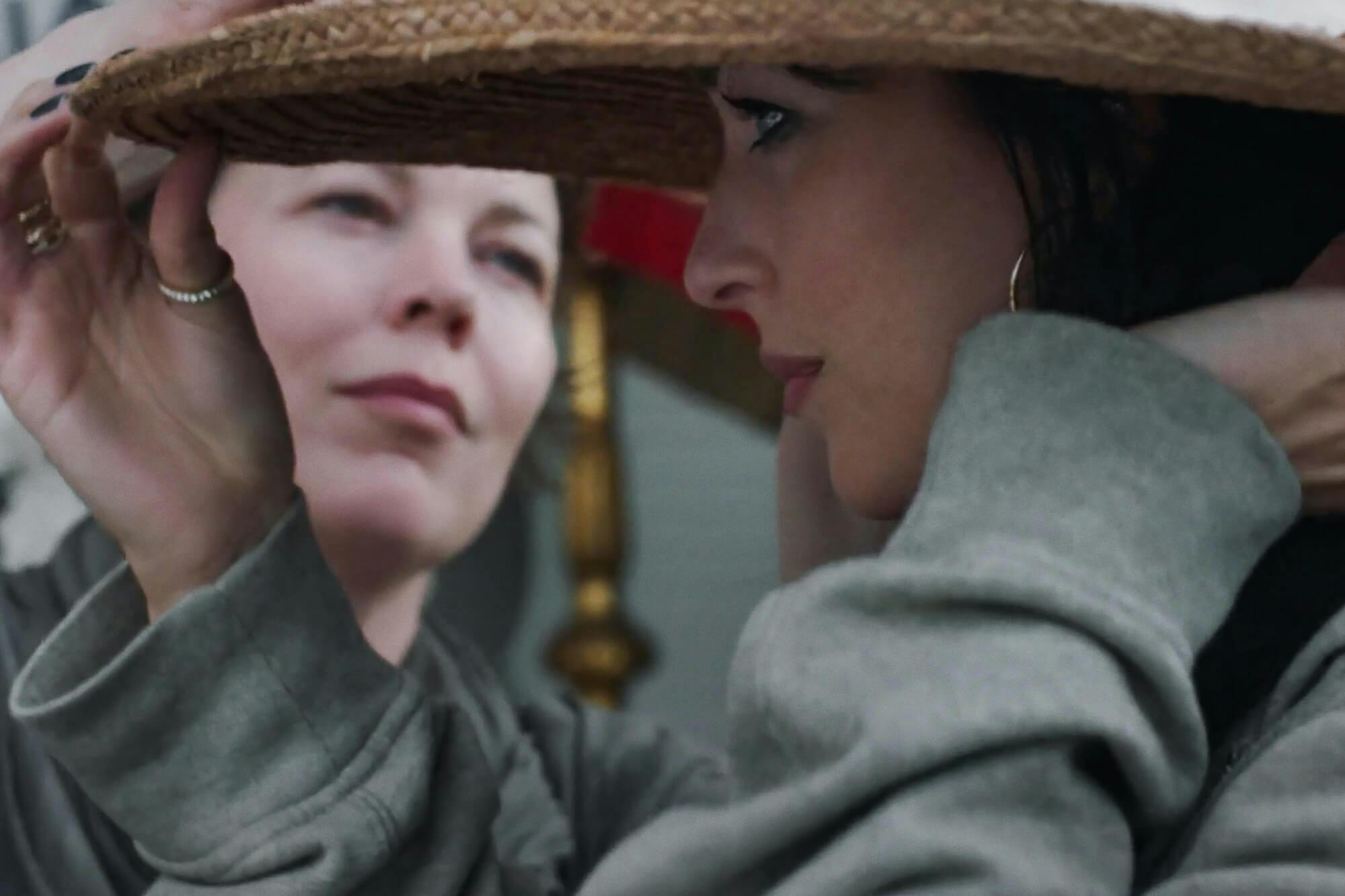 Leda (Olivia Colman) affixes a hat pin to Nina’s (Dakota Johnson) wide-brimmed hat in this close-up shot.