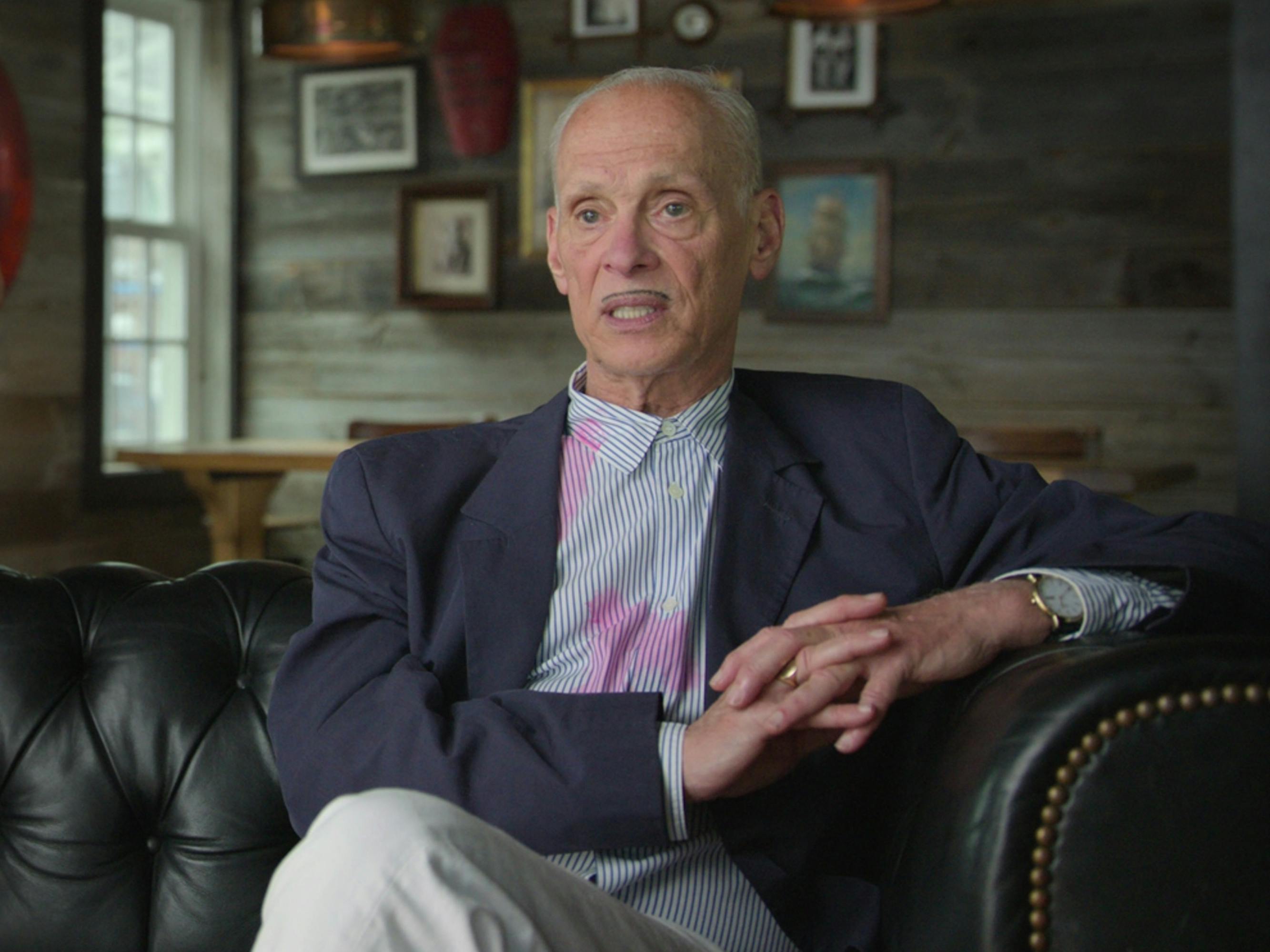 John Waters wears a blazer, color-less shirt and sits on a black leather couch.