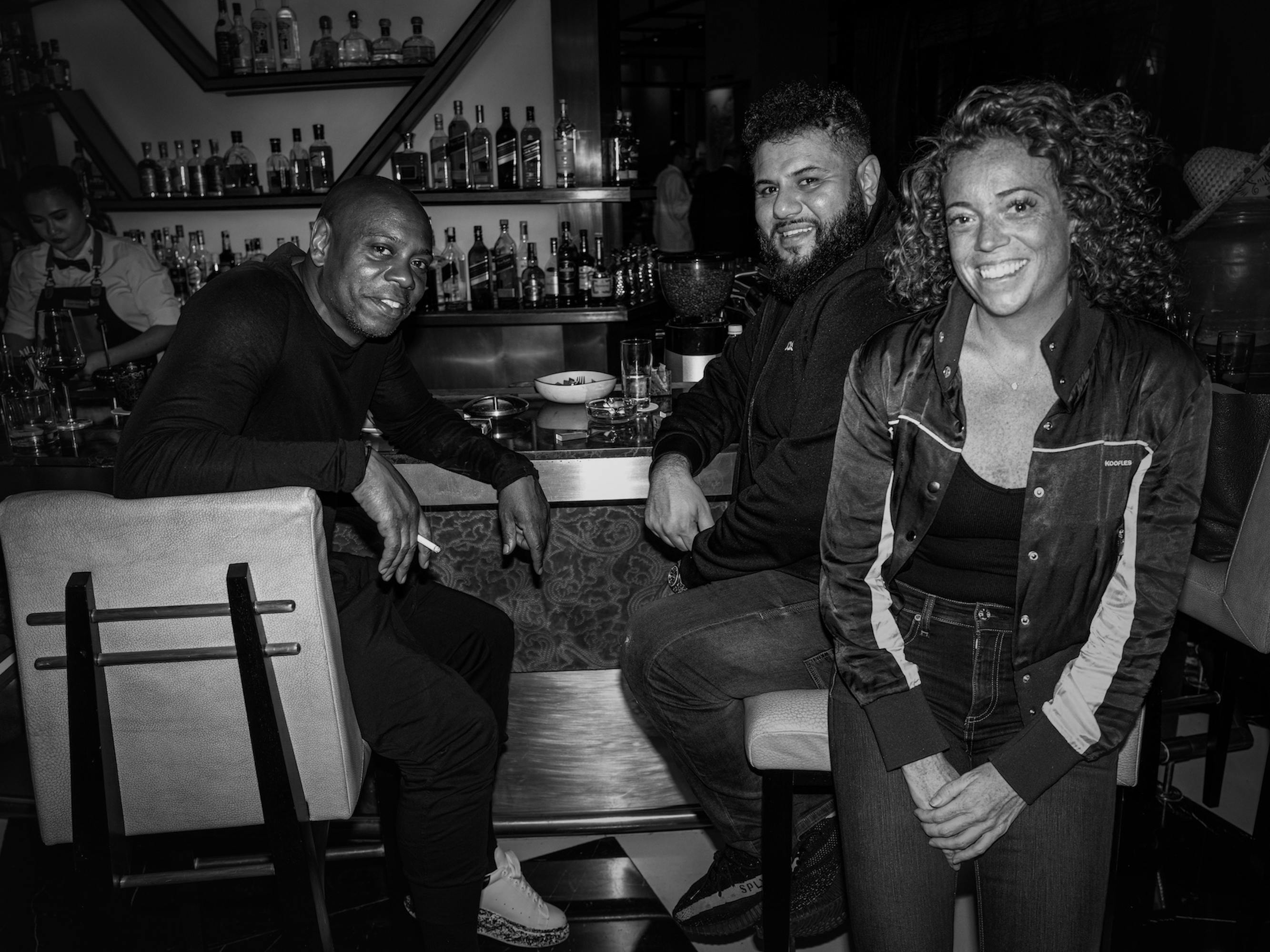 Chappelle, Mo Amer, and Michelle Wolf sit at a bar. They each wear all black and smile.