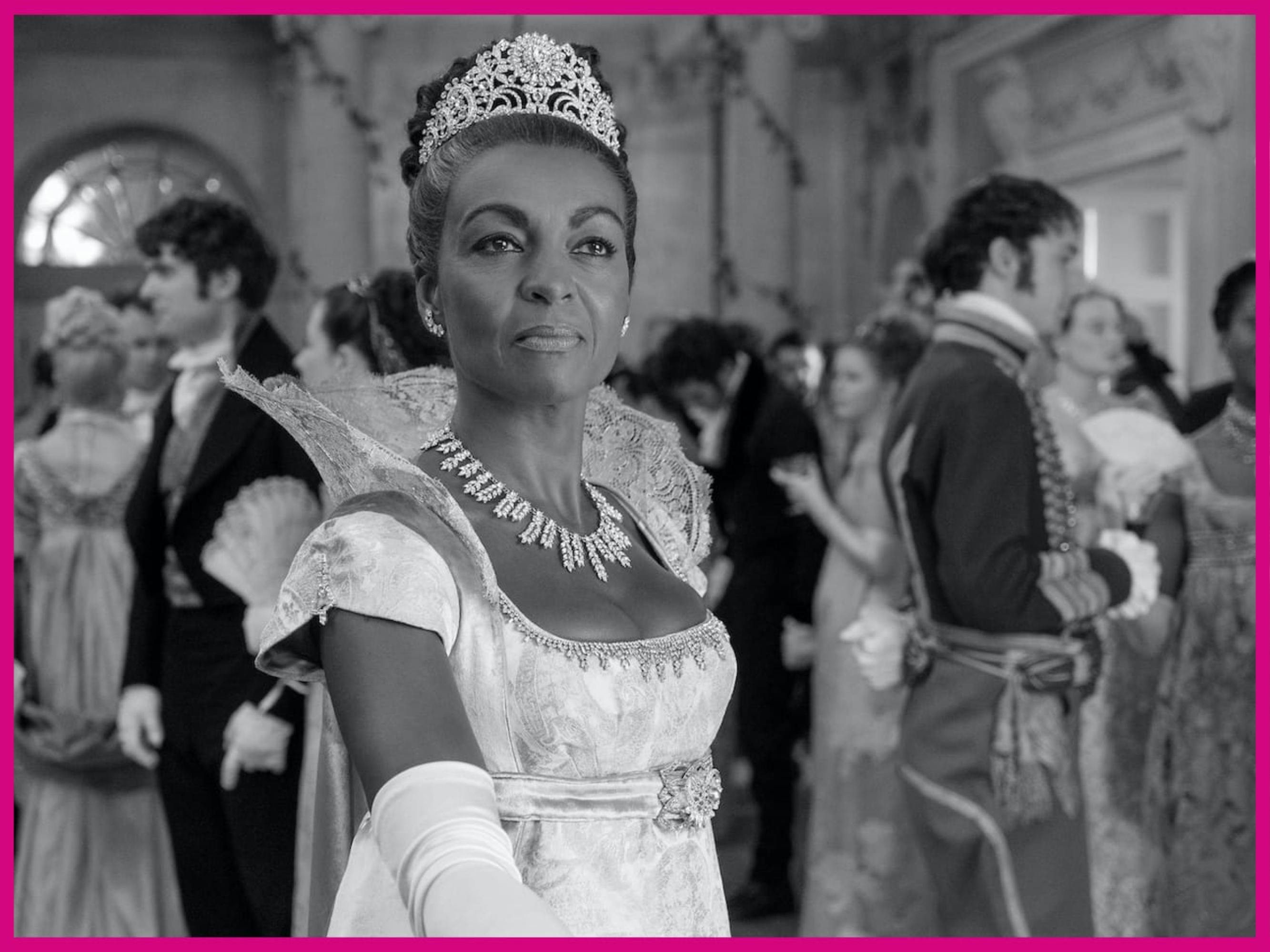 Lady Danbury (Adjoa Andoh) looks incredible in a silvery, white gown, complete with matching tiara and necklace. She reaches her hand out surrounded by other people at the ball dressed in similar finery.