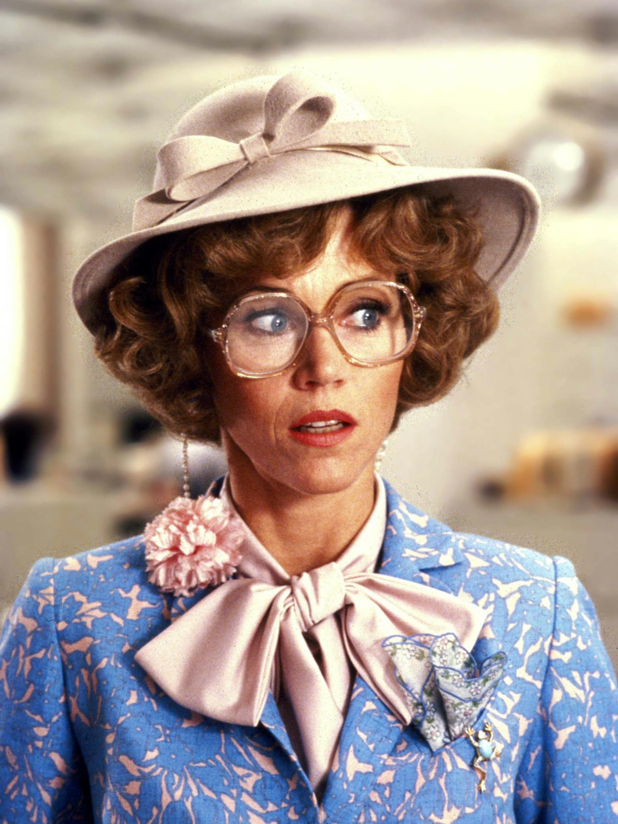 Jane Fonda looks surprised in a still from 9 to 5. She is wearing a blue and pink floral blazer, a pink pussy bow blouse, large clear-framed glasses, and a light pink hat atop her short curly hair.