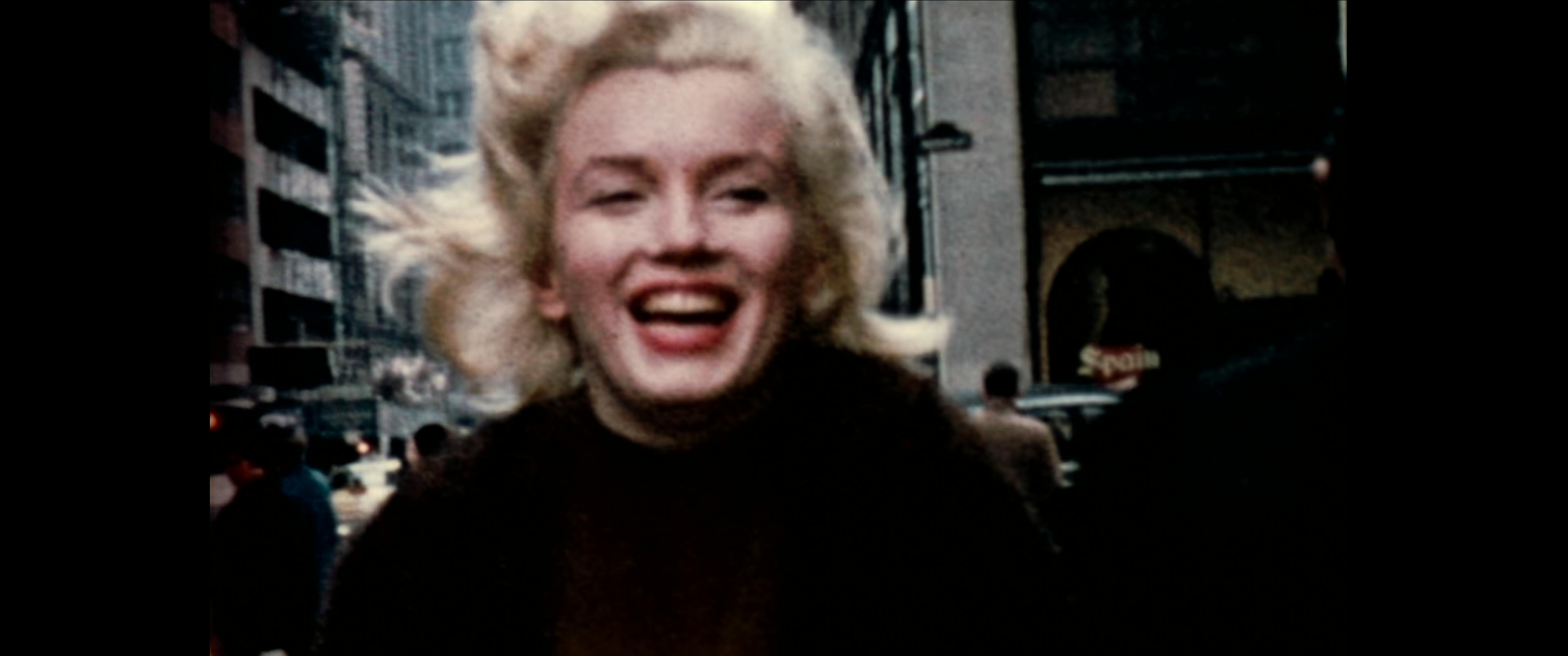 Marilyn Monroe wears a black top, her classic red lip, and smiles wide as she walks around what looks like a New York. 