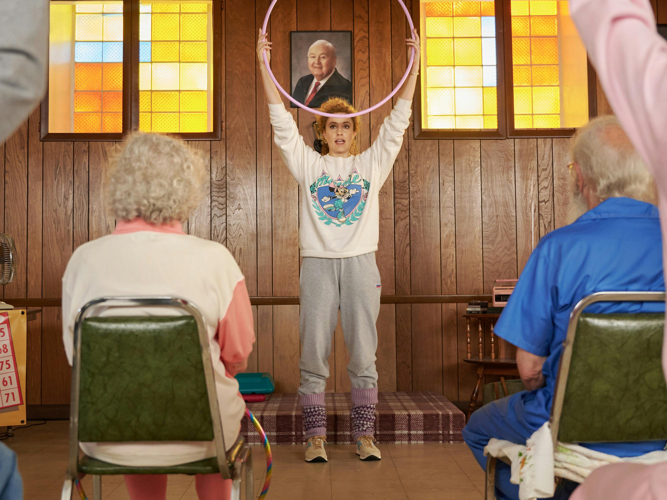 Greta Gerwig wears a white top and grey sweats and teaches an aerobics class to a bunch of grey-haired people.