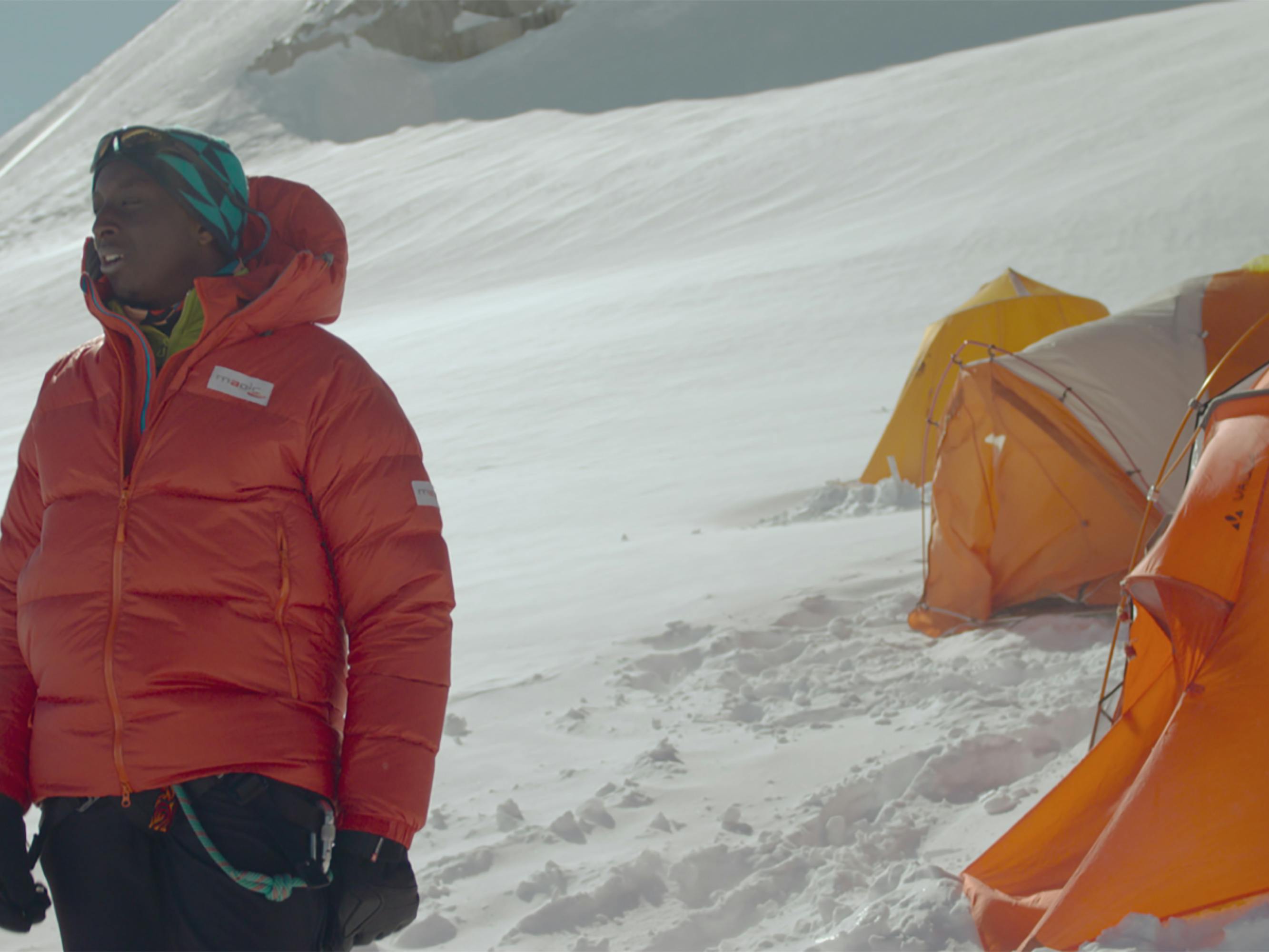 Samy (Ahmed Sylla) stands on a snowy expanse wearing an orange jacket and other hiking apparel. Behind him are orange tents.