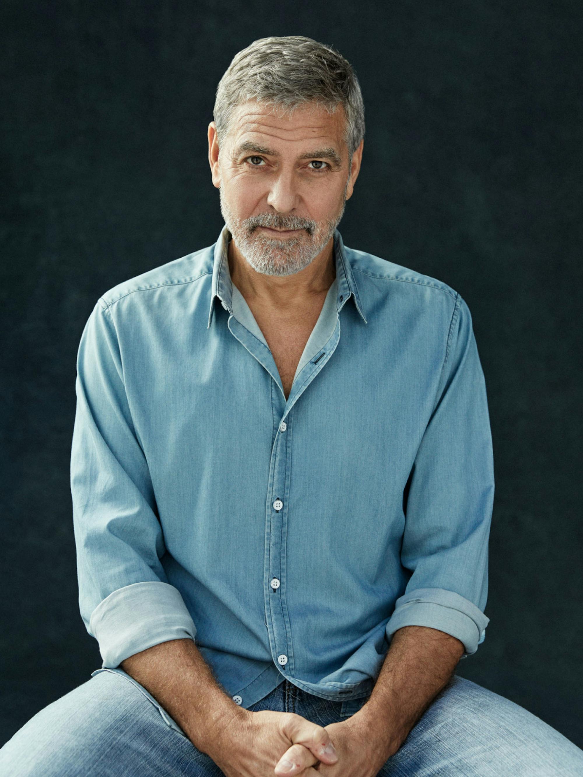 George Clooney in light-blue denim against a black background. With one look, he bores a hole right through the camera.