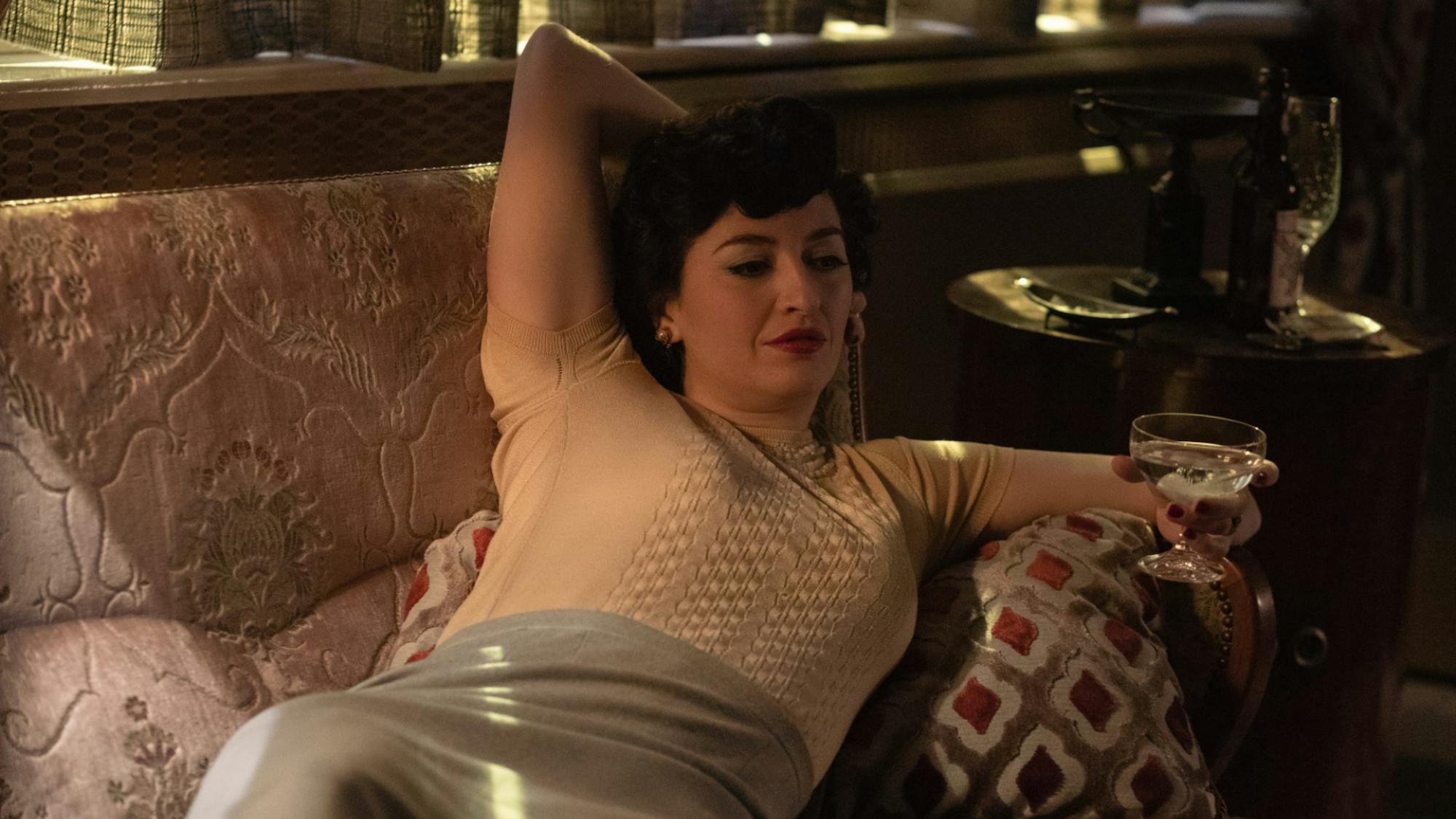 Alma Wheatley, played by Marielle Heller, relaxes on a multicolored couch drinking a cocktail. She wears a white shirt and a light blue latex skirt.