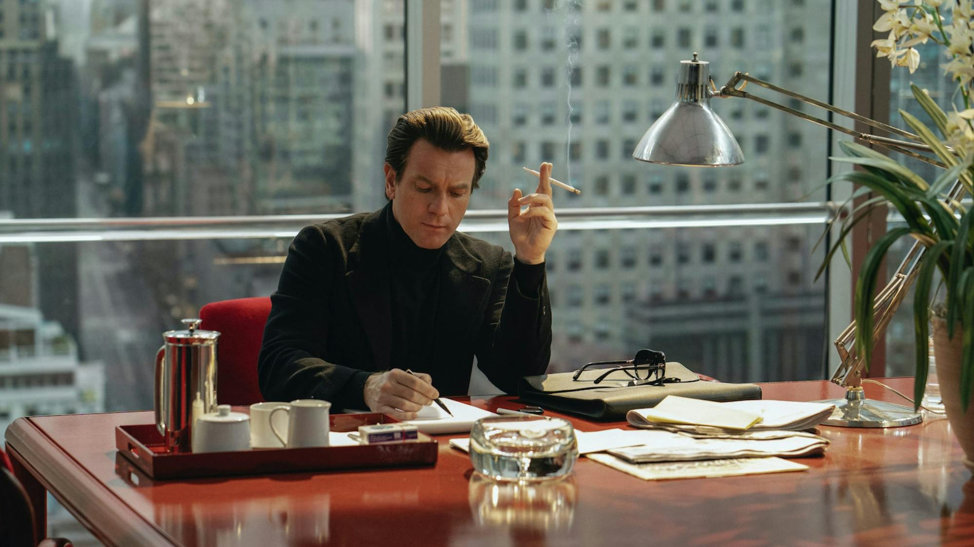 In this incredibly glamorous shot, Halston (McGregor) sits at a glossy cherry colored table, complete with red satiny chairs. A tray with what appears to be coffee or tea sits in front of him, as well as an ashtray, and papers strewn about. He wears an all black suit and, you guessed it, is smoking a cigarette! Behind him are completely unobstructed windows that show buildings and a busy city scene beneath. 