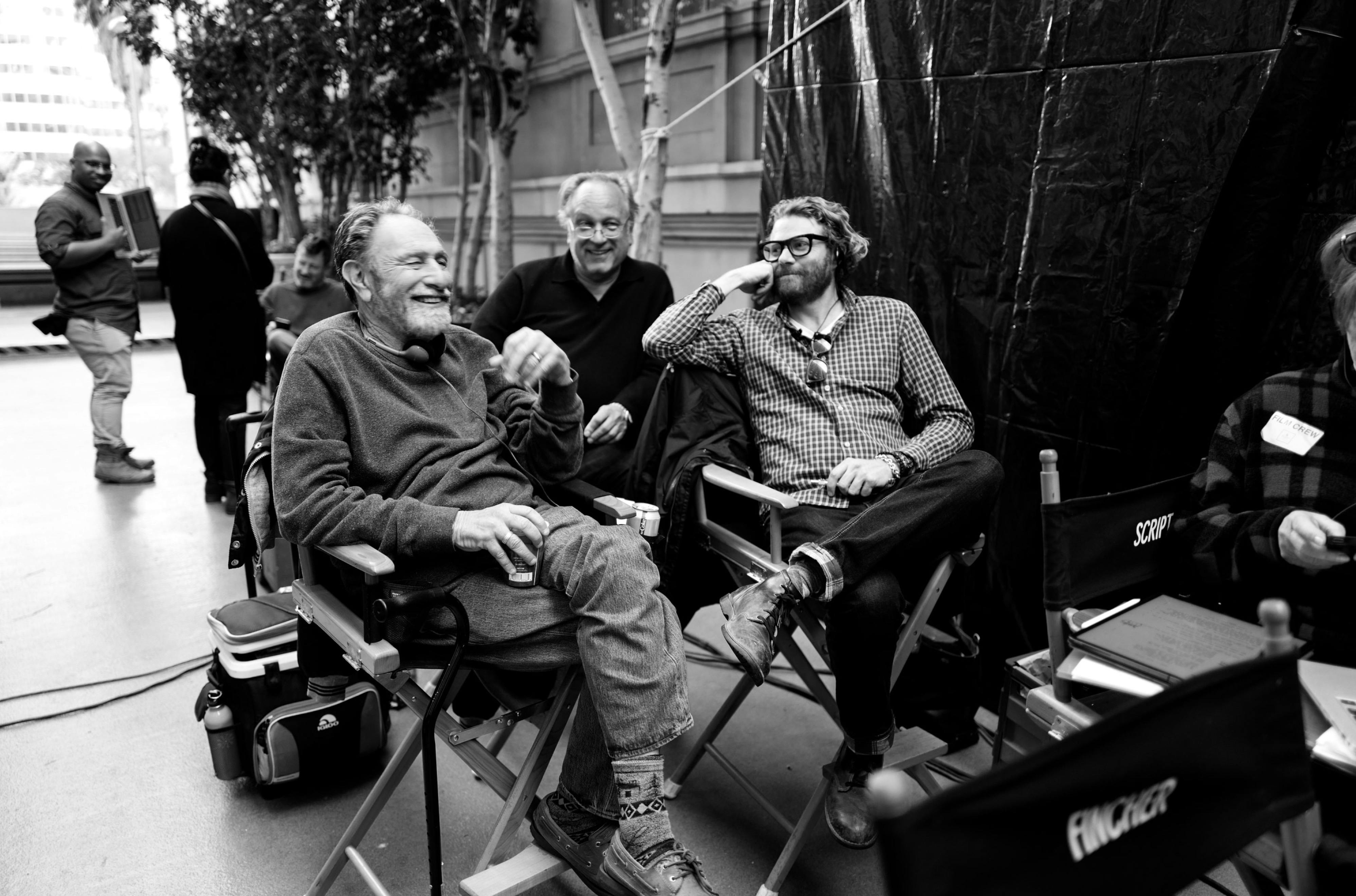 A black-and-white behind-the-scenes photo of Roth with Urbanski and Messerschmidt on the set of Mank. Roth and Fincher are seated in director’s chairs, smiling.