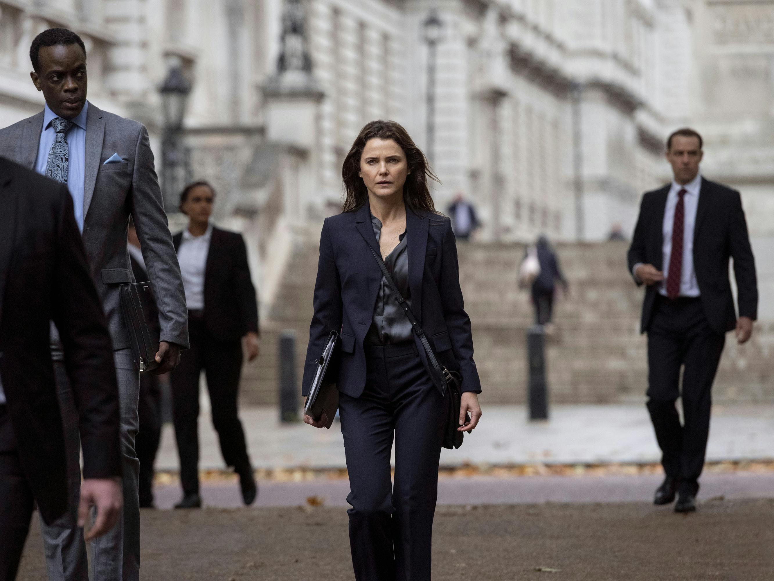 Stuart Heyford (Ato Essandoh) and Kate Wyler (Keri Russell) walk on a city street looking intense, surrounded by other intense-looking people in suits.