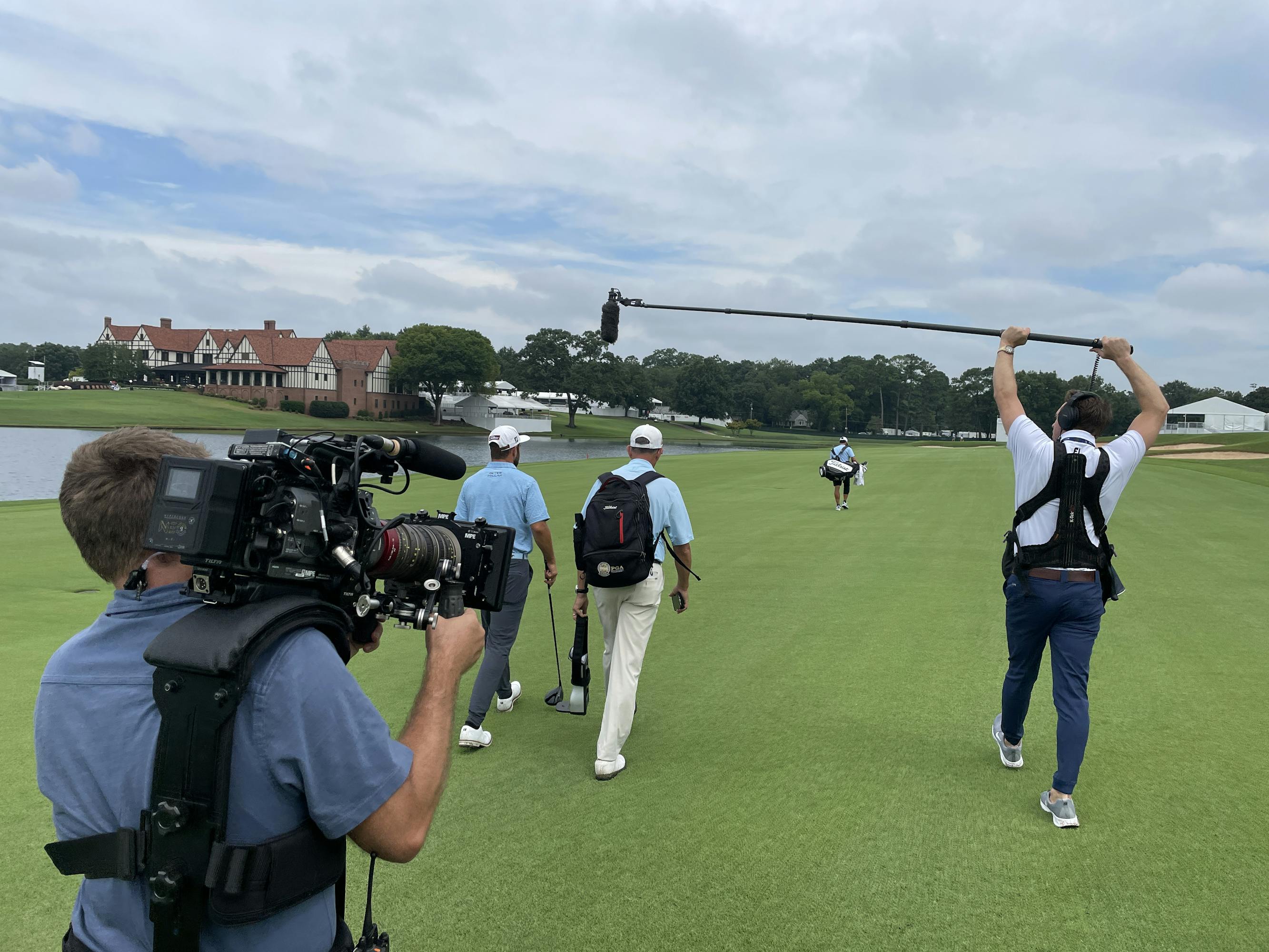A golfer and his caddie walk along the green, trailed by a cameraman and a boom operator.