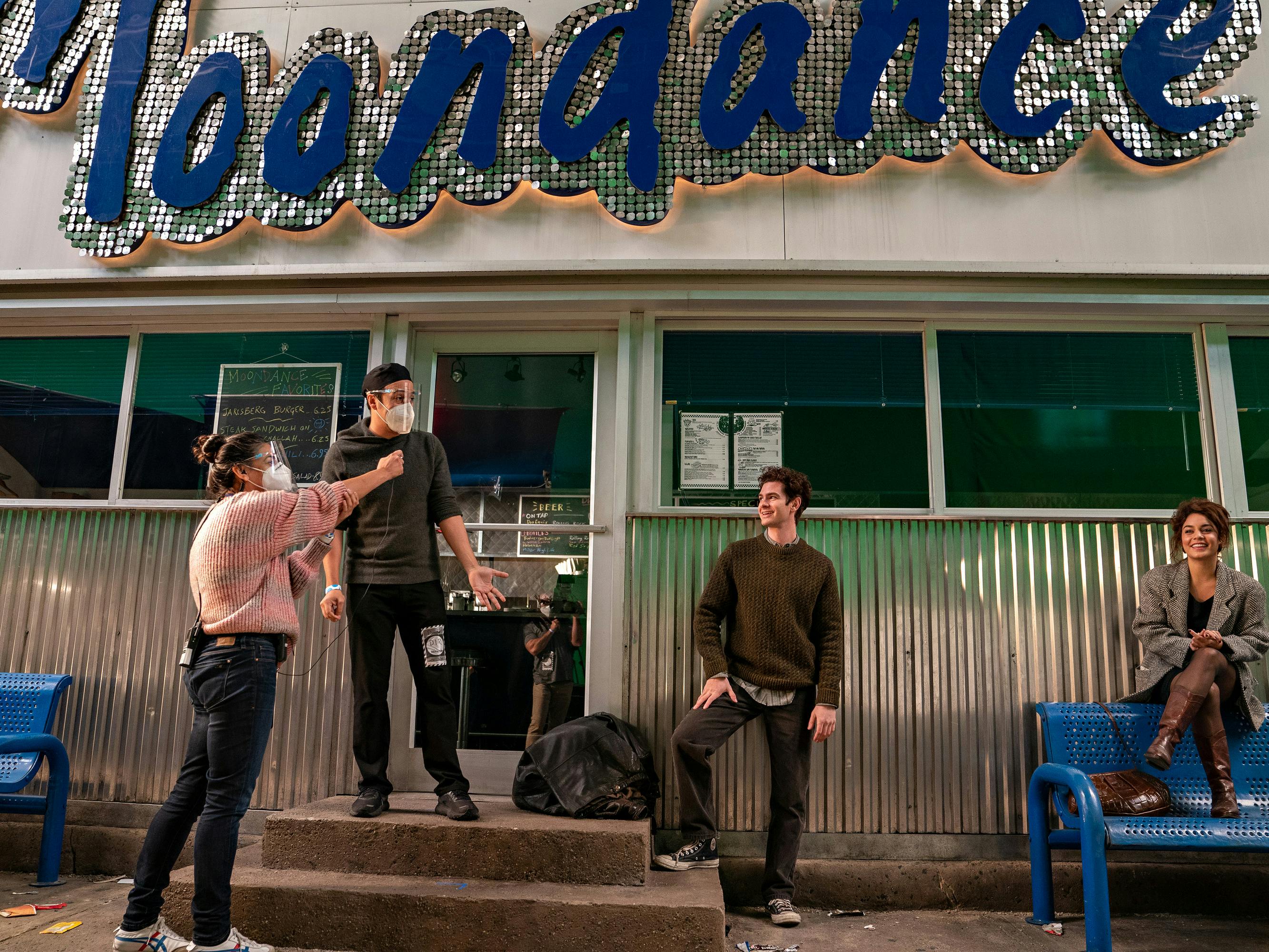 First Assistant Director Mariela Comitini, Producer-director Lin-Manuel Miranda, Andrew Garfield, and Vanessa Hudgens, and Firstthe tick, tick . . . BOOM! crew member stand outside the Moondance Diner. The crew is masked, Garfield wears a green sweater, and Hudgens wears brown boots and an oversized blazer.