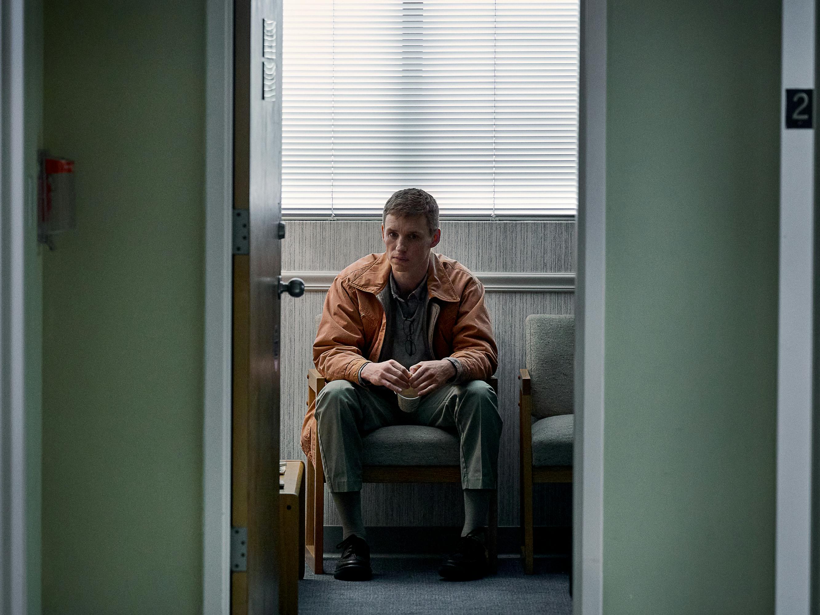 Charles Cullen (Eddie Redmayne) wears an orange coat, green pants, and sits in a waiting room.