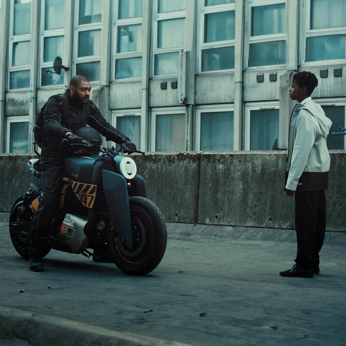 Izi (Kane Robinson) and Benji (Jedaiah Bannerman) meet on a concrete bridge. Izi is on a motorcycle. Behind them are weathered apartment buildings. 