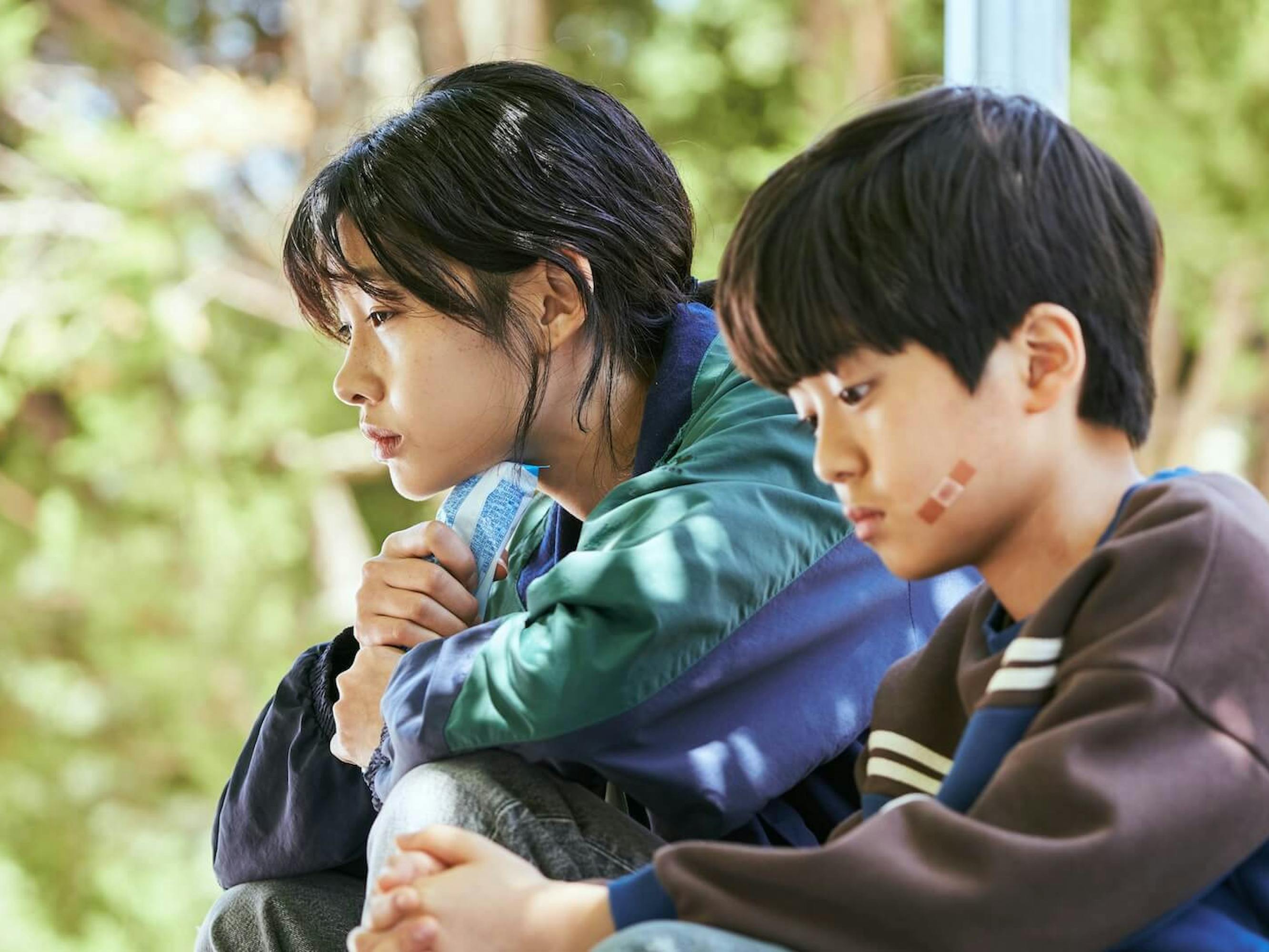 Sae-byeok (Jung Ho-yeon) and her younger brother sit outside in sun-dappled light. She wears a blue-and-green jacket and he wears a brown sweater with a small Band-Aid on his face.