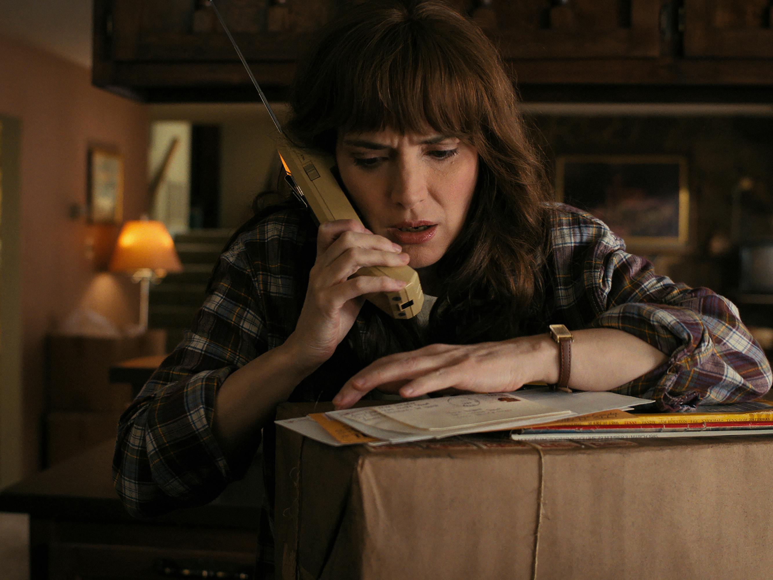Joyce Byers (Winona Ryder) talks into a phone as she rests on a large package. She wears a flannel shirt and has impressive bangs.