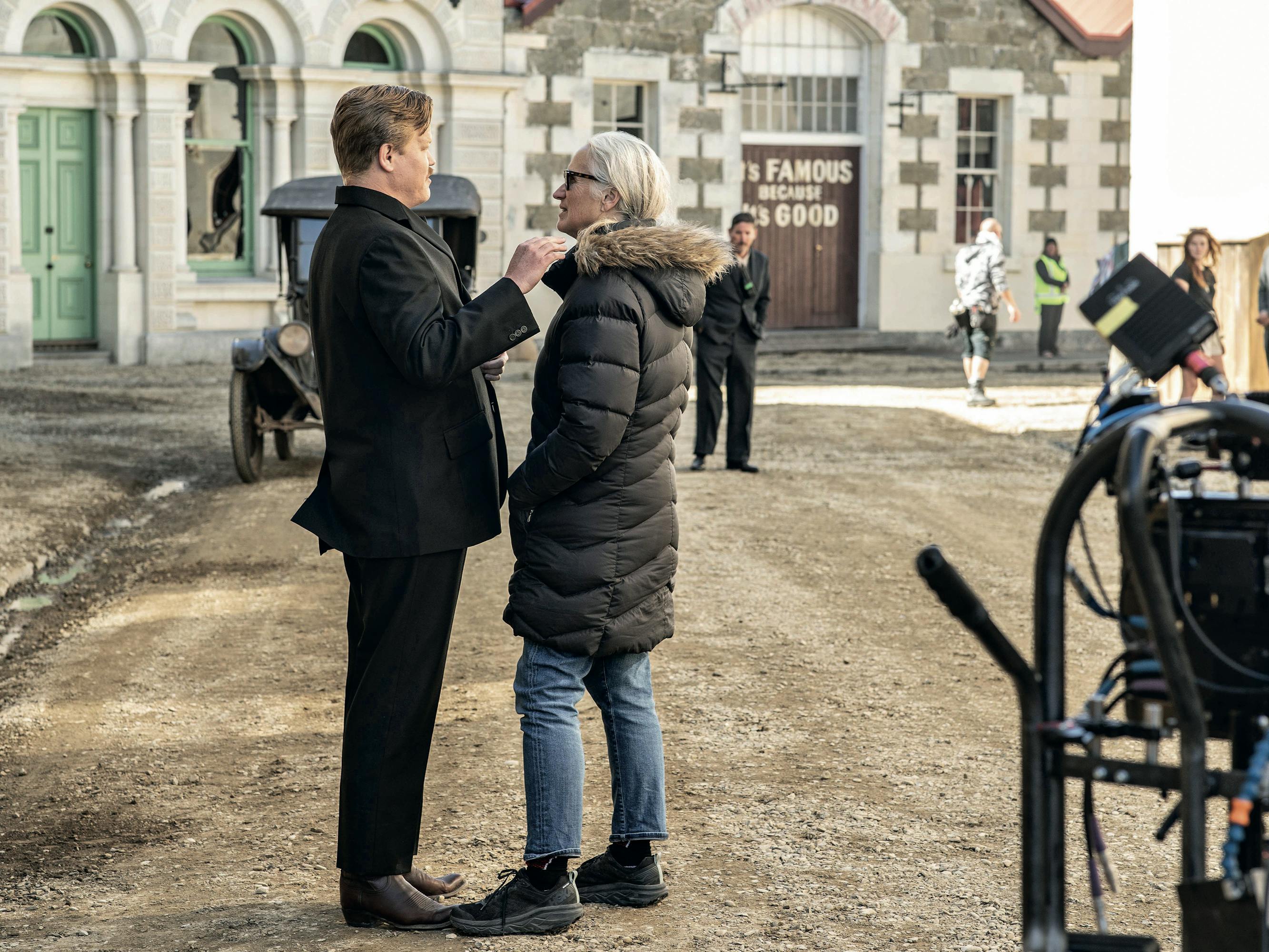 Jesse Plemons and Jane Campion stand in the middle of a dusty set. There is equipment in the bottom left corner of the shot.