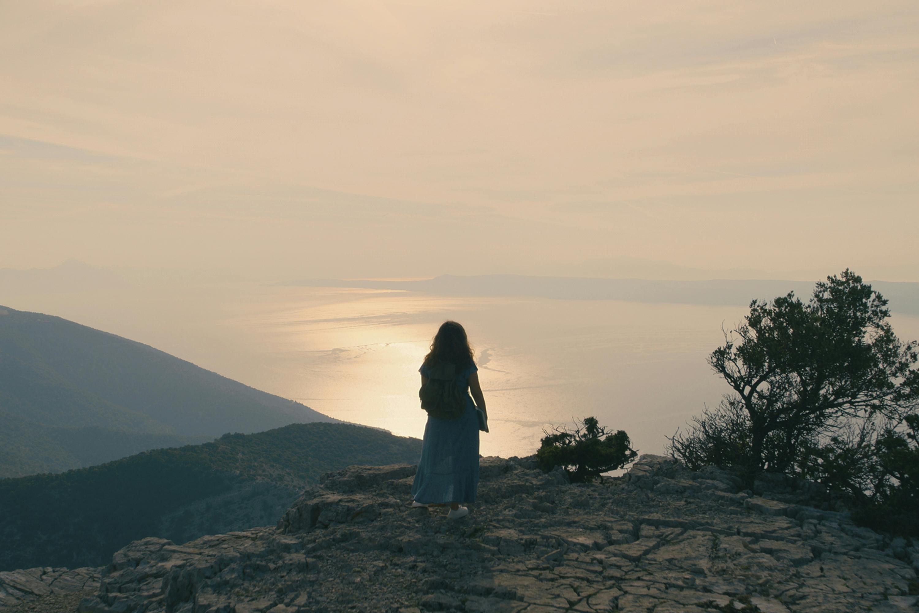 Zeynep (Naomi Krauss) stands on a cliff’s edge overlooking a beautiful sun setting on the still water.
