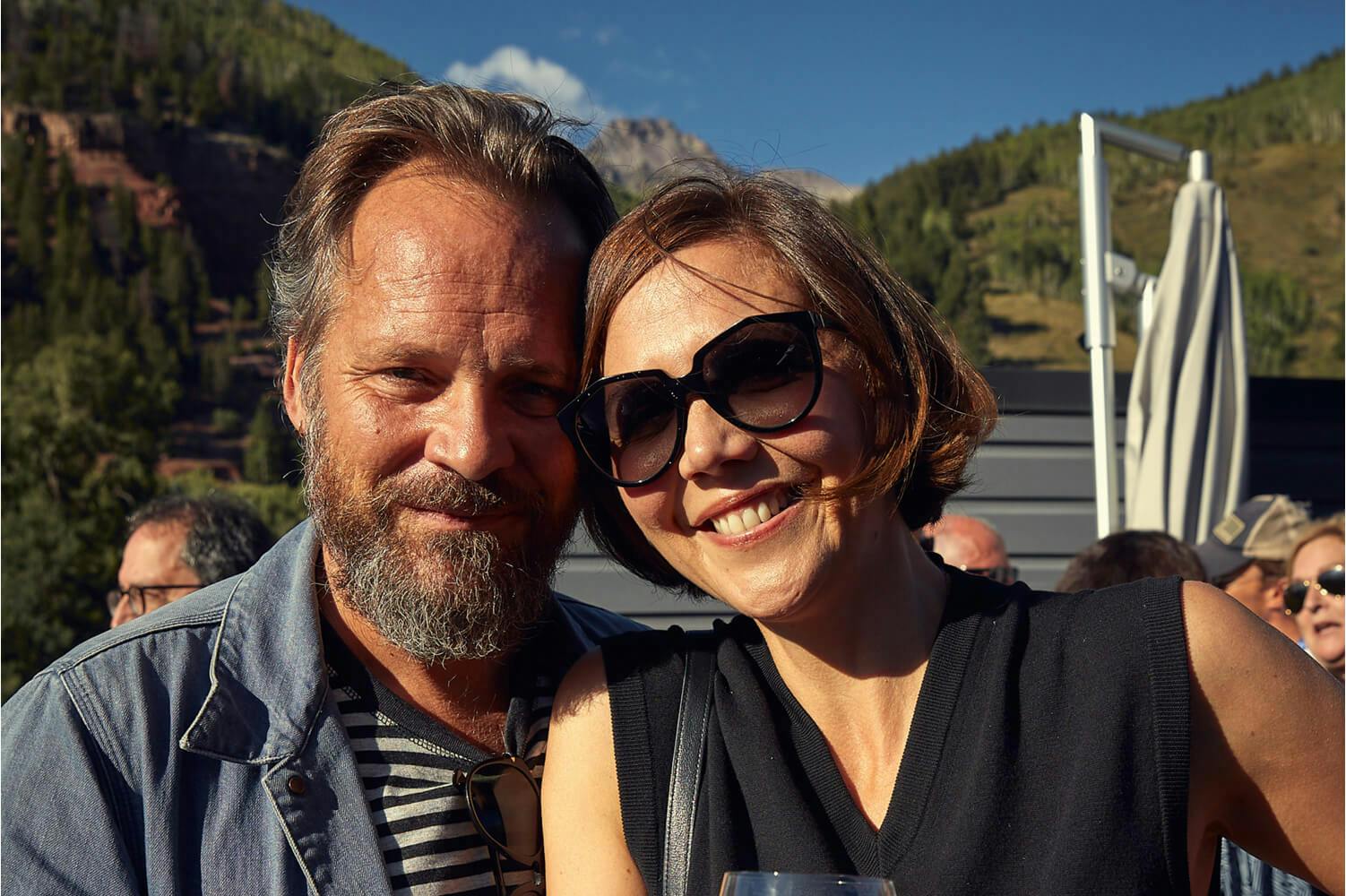Maggie Gyllenhaal and Peter Sarsgaard at the Telluride Film Festival, 2021 wearing casual attire while standing at a rooftop event with the Colorado mountains in the background.