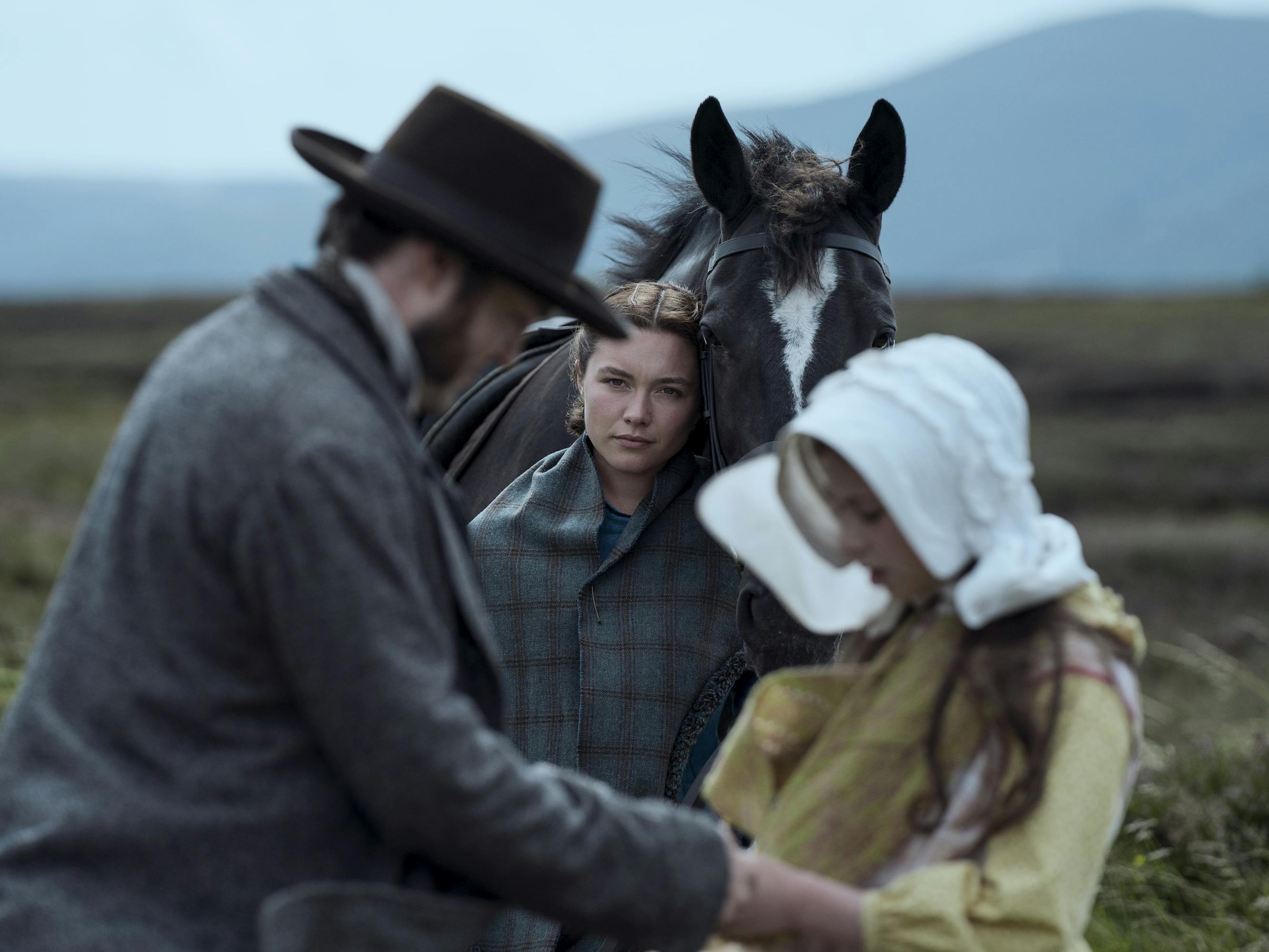 Tom Burke, Florence Pugh, and Kíla Lord Cassidy stand in a windy landscape.