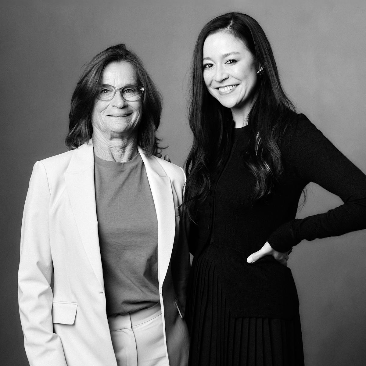 Bonnie Stoll and Elizabeth Chai Vasarhelyi stand together against a grey wall. Bonnie wears a white suit and Elizabeth wears all black.