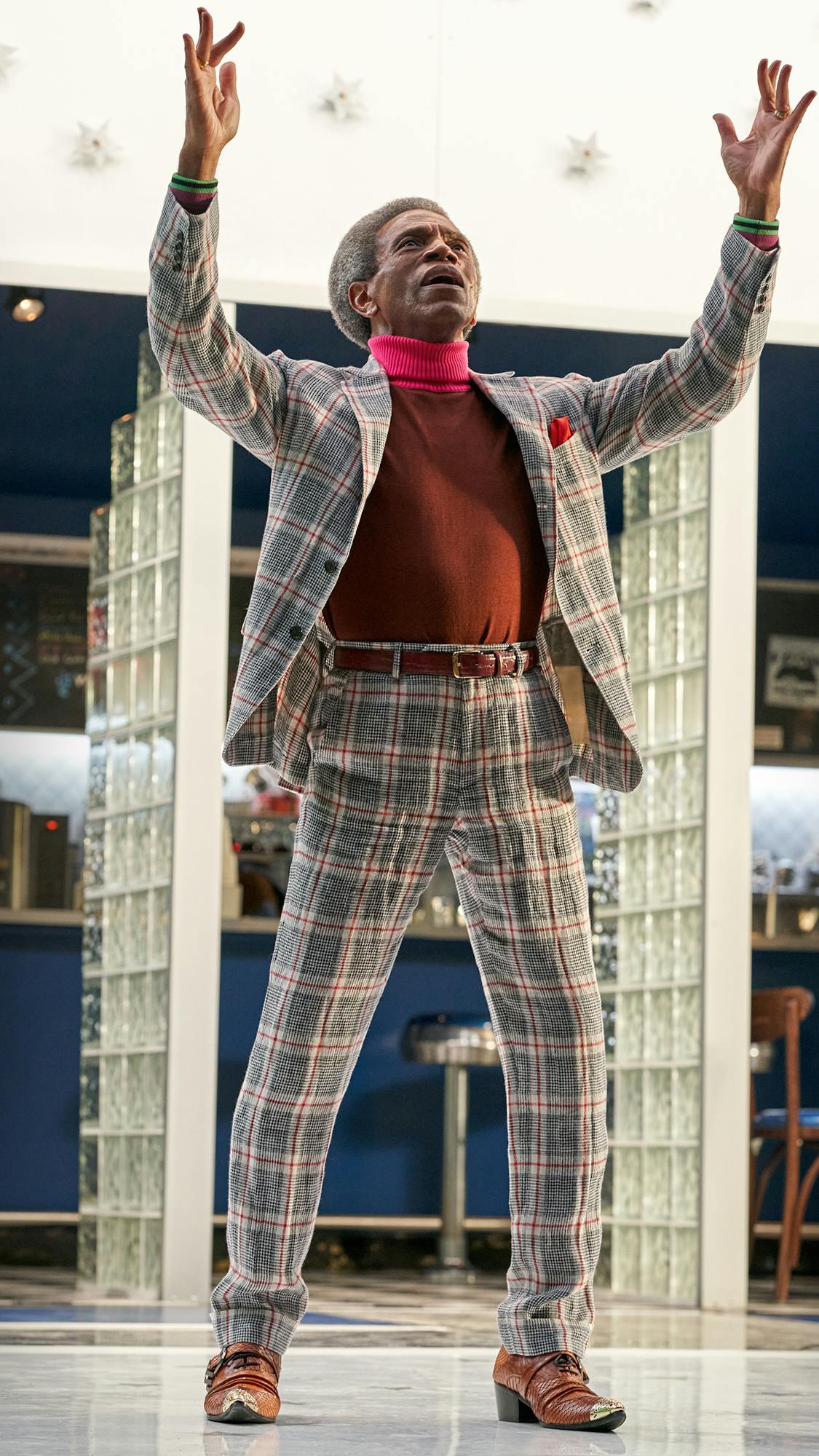 André De Shields wears a plaid suit and a hot pink turtle neck and stands outside the Moondance Diner.