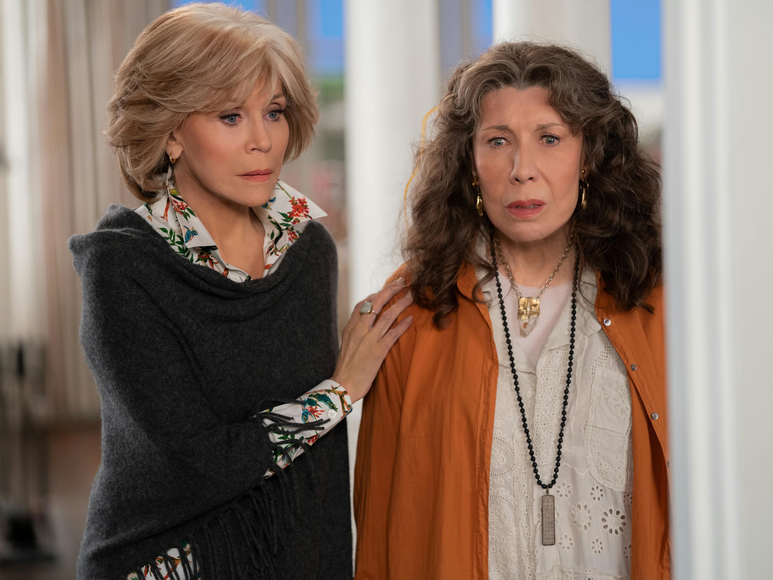 Jane Fonda and Lily Tomlin look alarmed and stand by a door frame. Fonda wears a black sweater and pattered shirt beneath, Tomlin wears an orange cardigan.