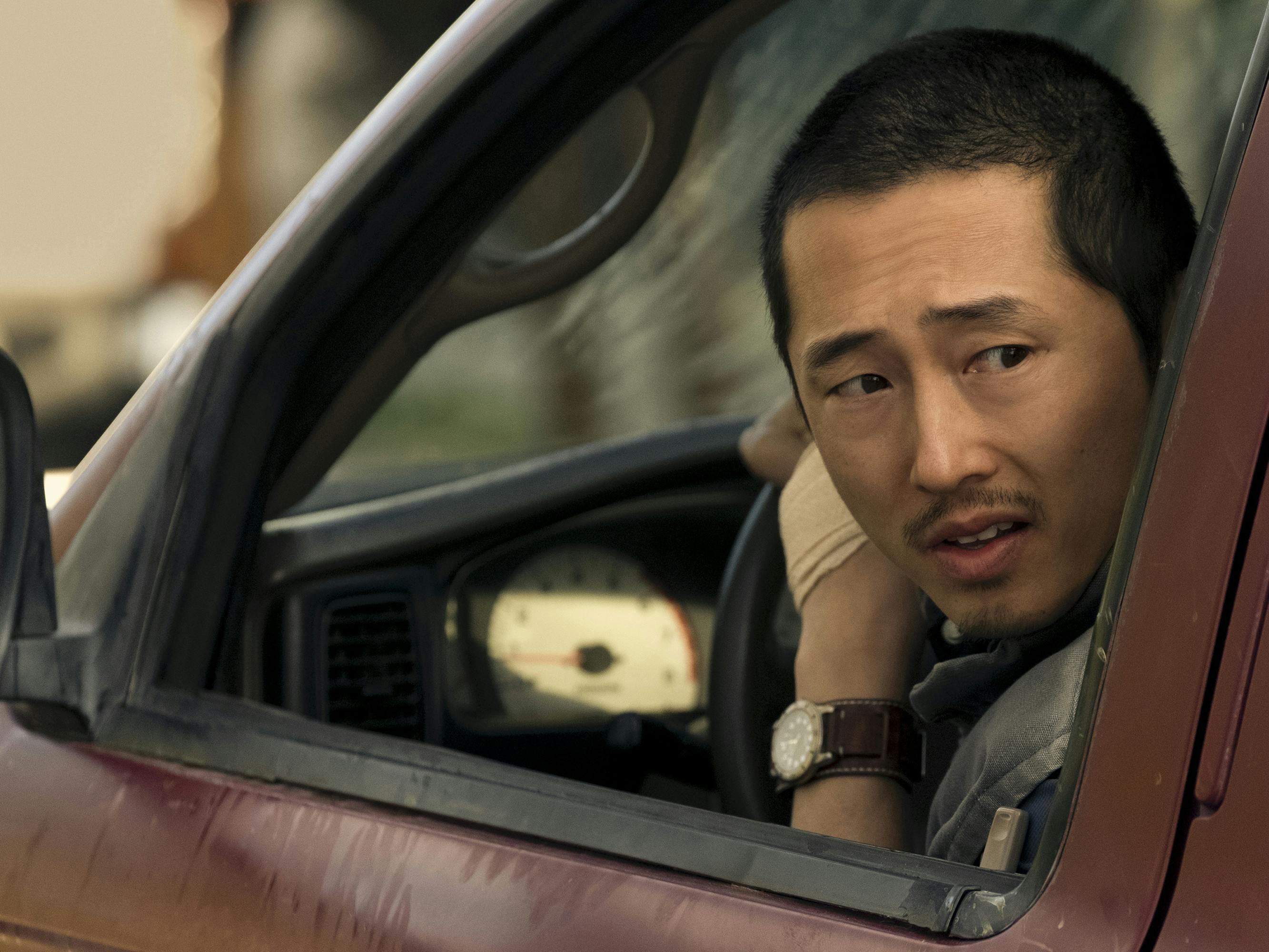 Steven Yeun drives his red pickup truck, looking behind him out the window. 