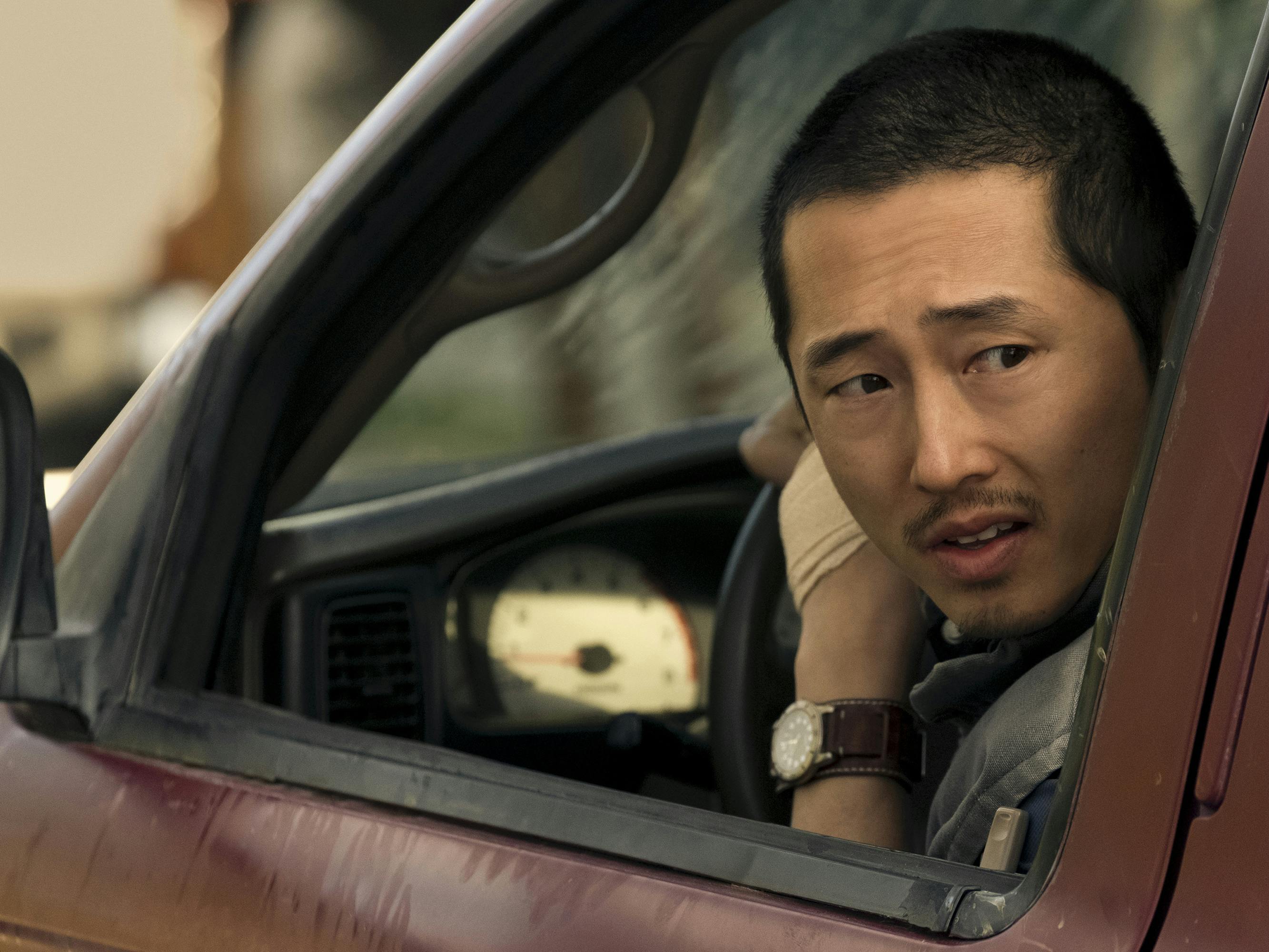 Steven Yeun drives his red pickup truck, looking behind him out the window.