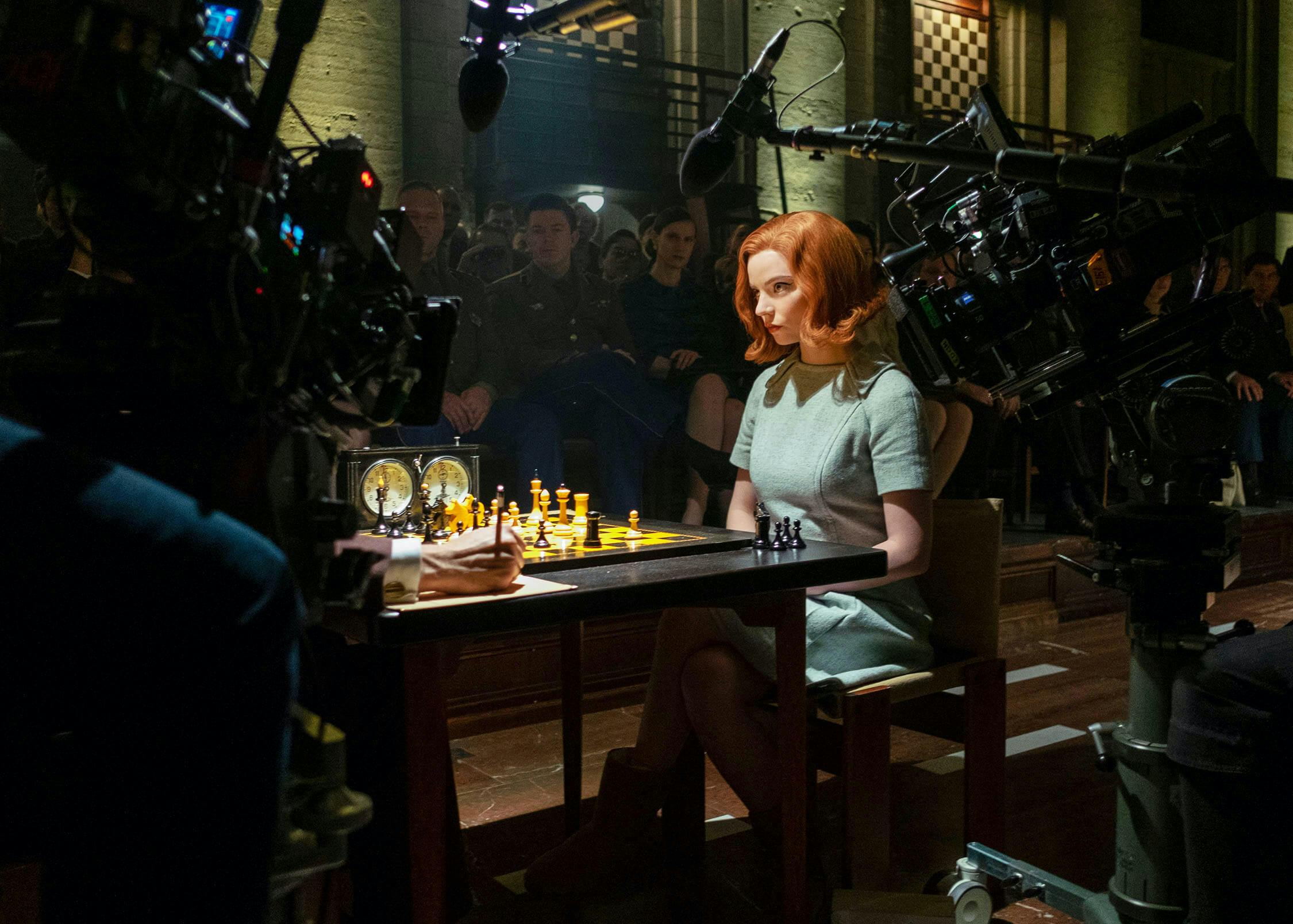 Beth Harmon (Anya Taylor-Joy) sits in an open hall. The scene is filled with camera gear and actors watching from the background. She wears a blue set and her bright red hair stands out in the darkness.